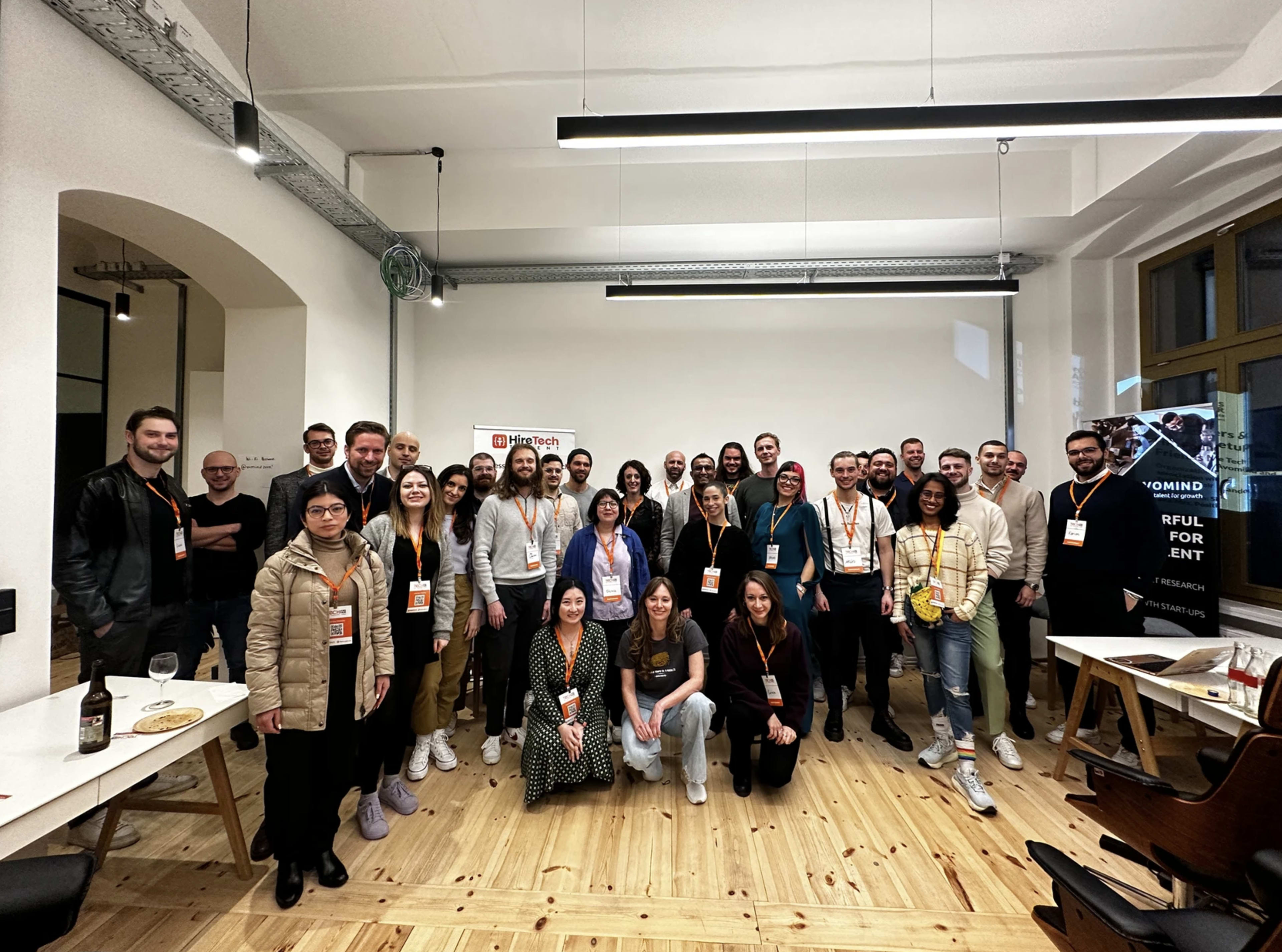A large group of people stands together in a well-lit room, smiling at the camera, with a mix of casual and business attire.