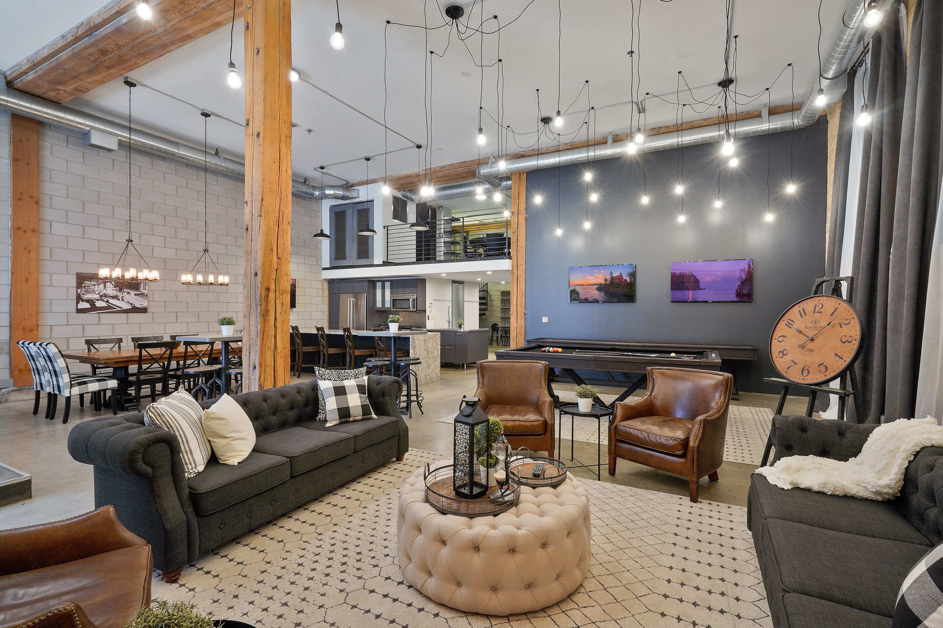 A modern open-concept living space with a combination of seating areas, a dining area, and a kitchen, featuring industrial-style decor and hanging light fixtures.