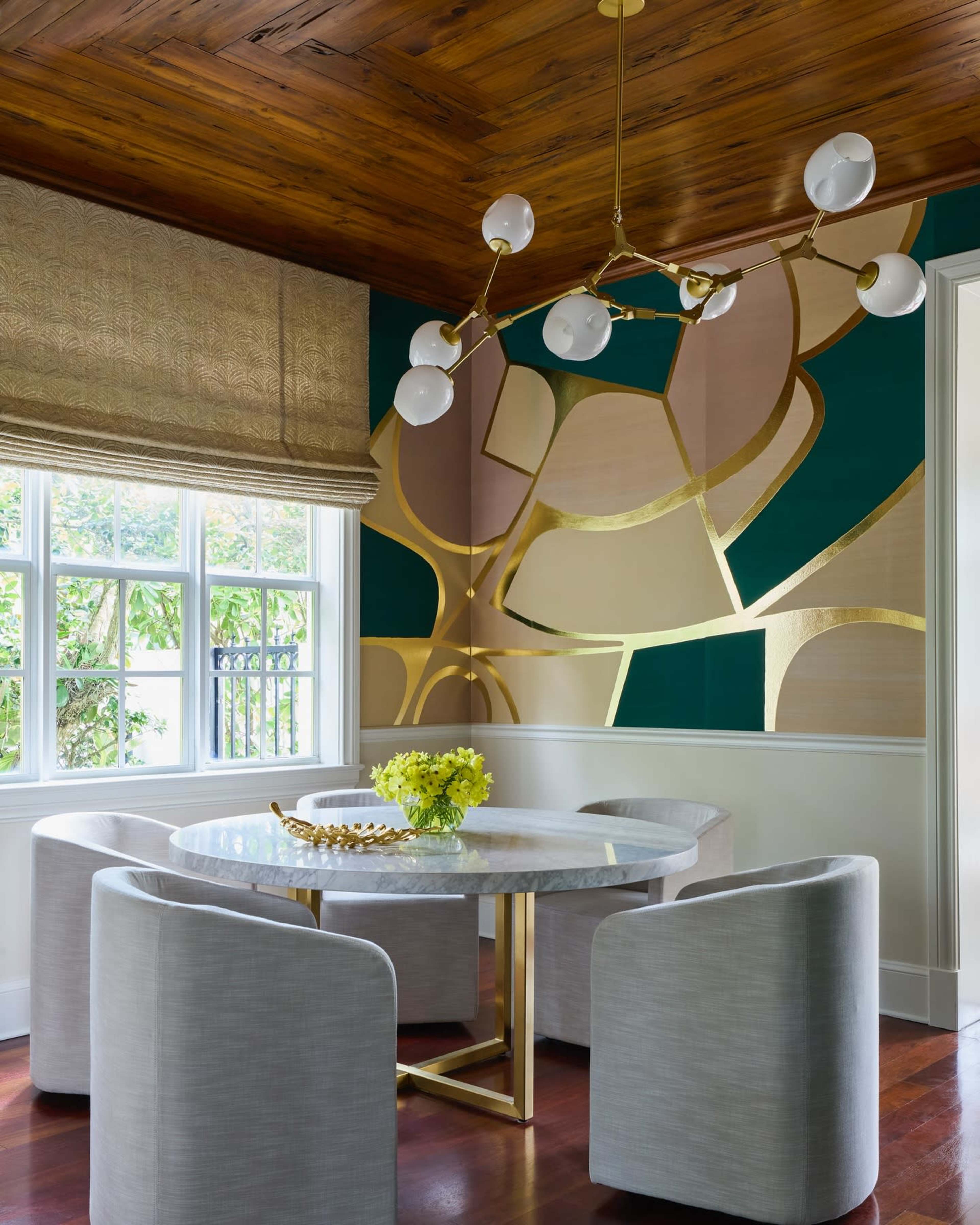 The image shows a dining area with a round marble table surrounded by four light-colored upholstered chairs, a modern chandelier overhead, and a wall featuring abstract geometric patterns in green, gold, and beige.