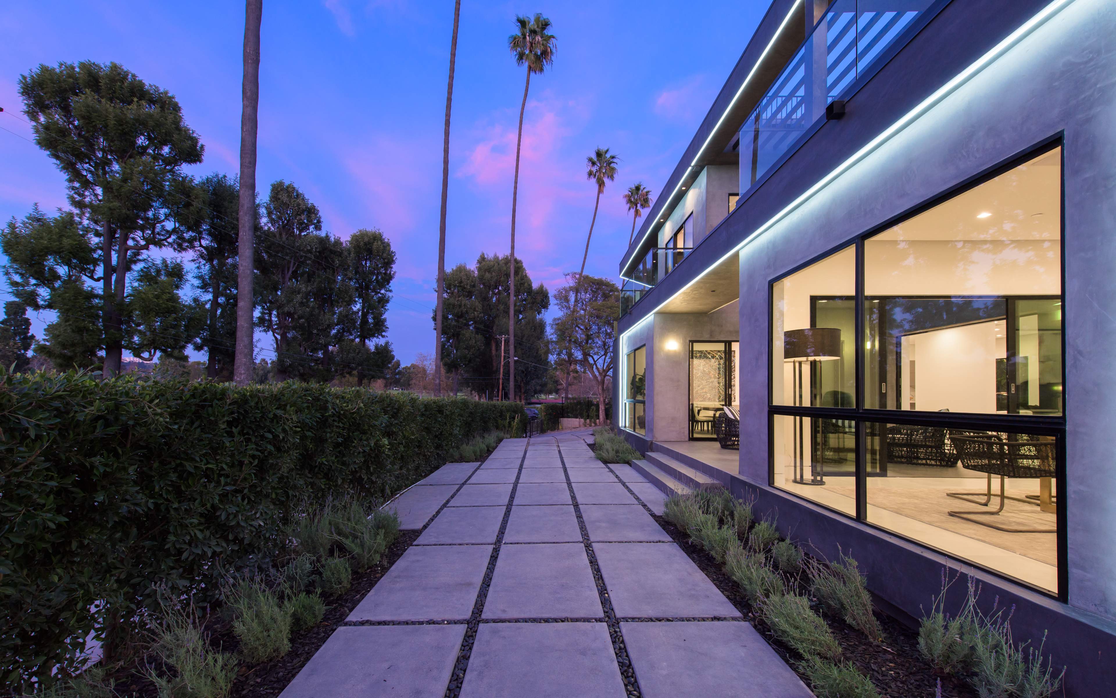 A modern home with large glass windows is framed by a concrete walkway and surrounded by palm trees at dusk.