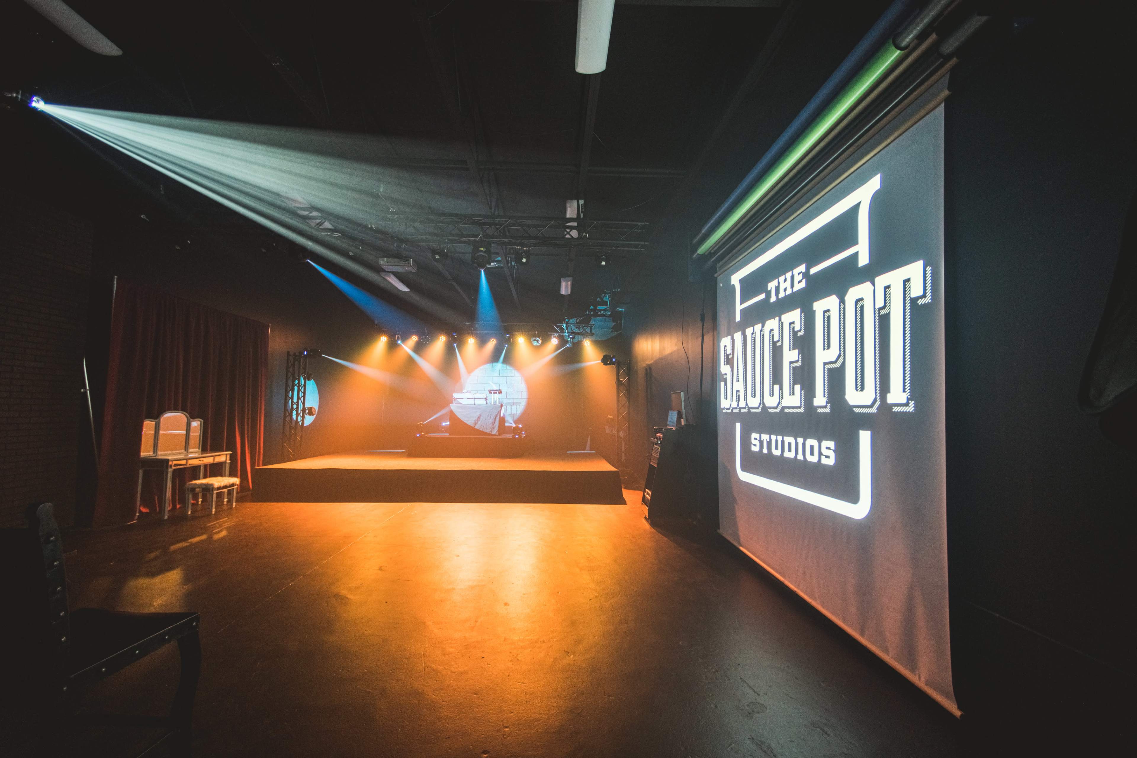 An interior of a performance studio called "The Sauce Pot Studios," featuring a stage with lighting and a large logo displayed on a screen.
