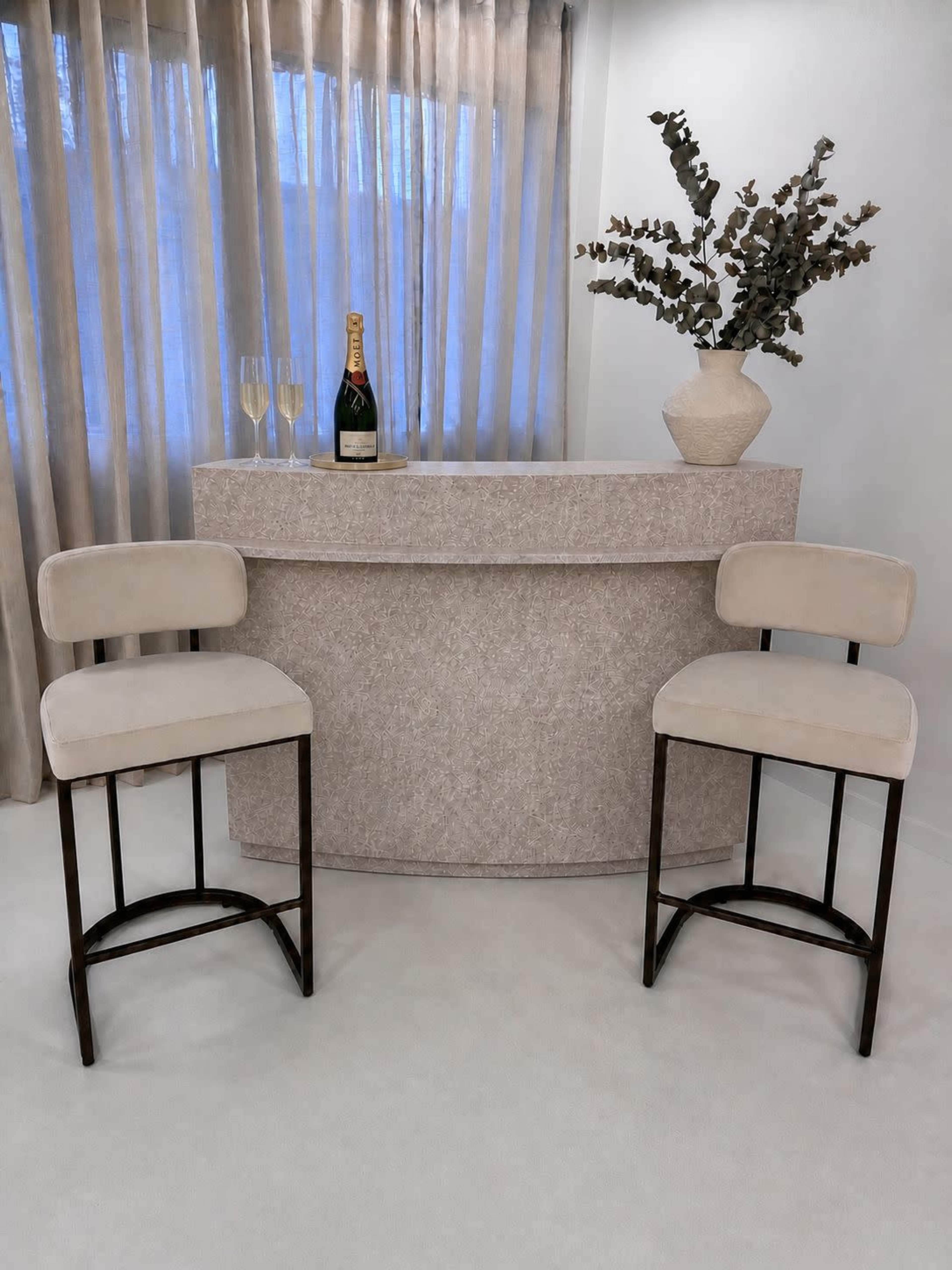 A simple bar area features two upholstered stools beside a rounded, textured bar, with a bottle of champagne and glasses on the counter, along with a decorative vase and greenery.