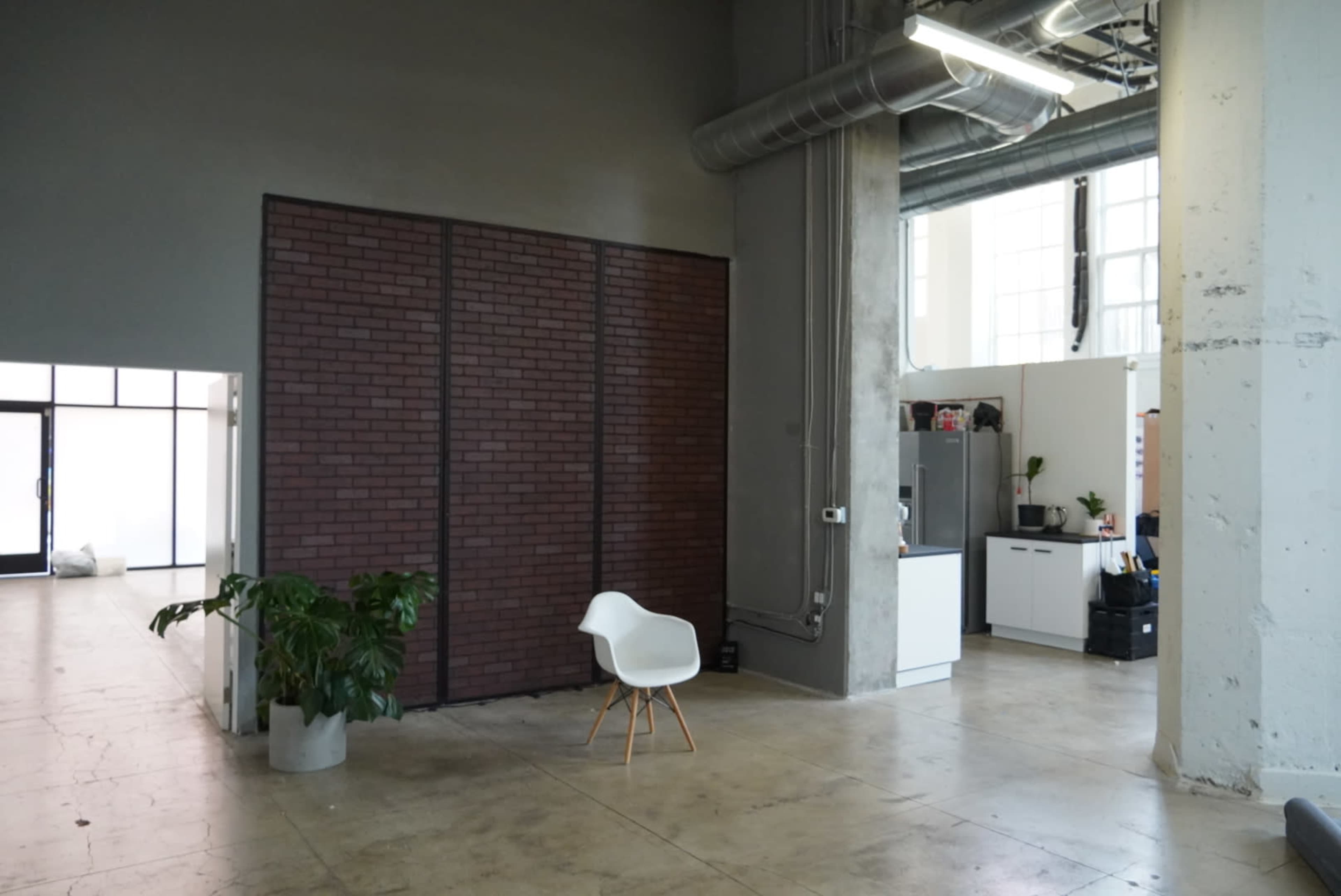 An open office space featuring a gray wall with a brick accent, a white chair on a wooden stand, a leafy plant, and a kitchenette area in the background.