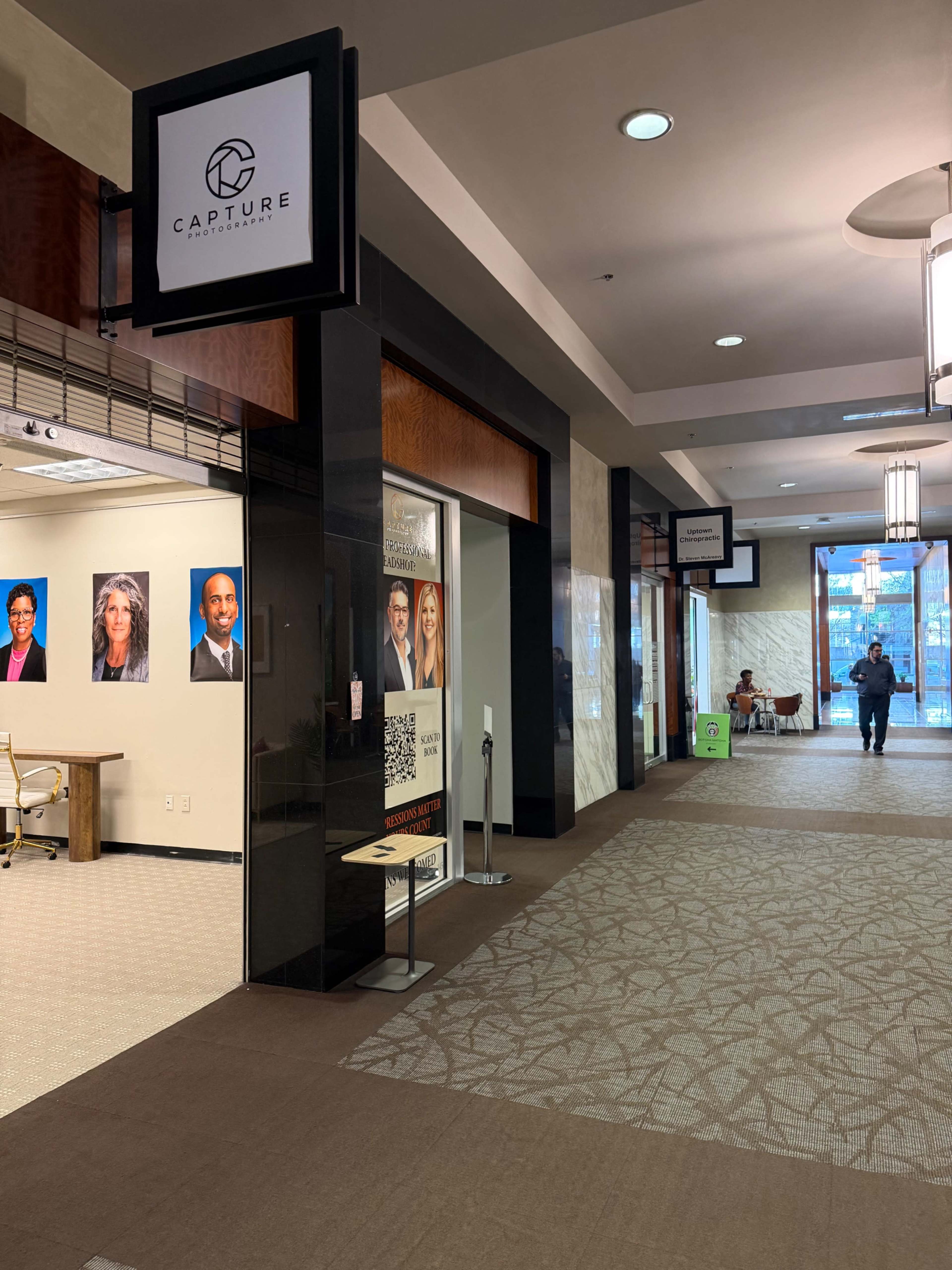 The image shows a corridor of a modern building with various shops and offices, featuring a sign for "Capture" and several framed portraits on the wall.