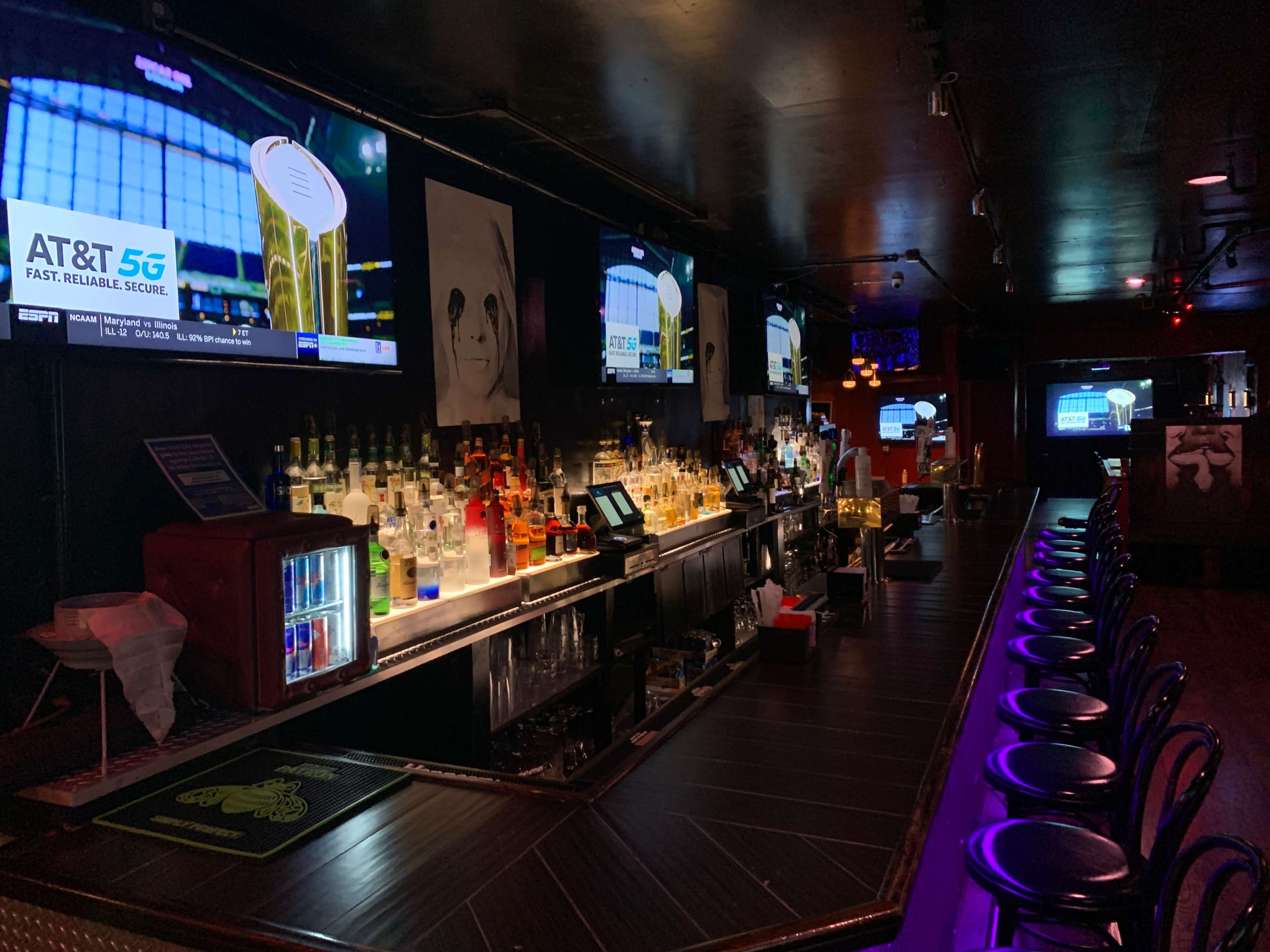 A dimly lit bar with a long counter, adorned with various liquor bottles and multiple televisions displaying sports.