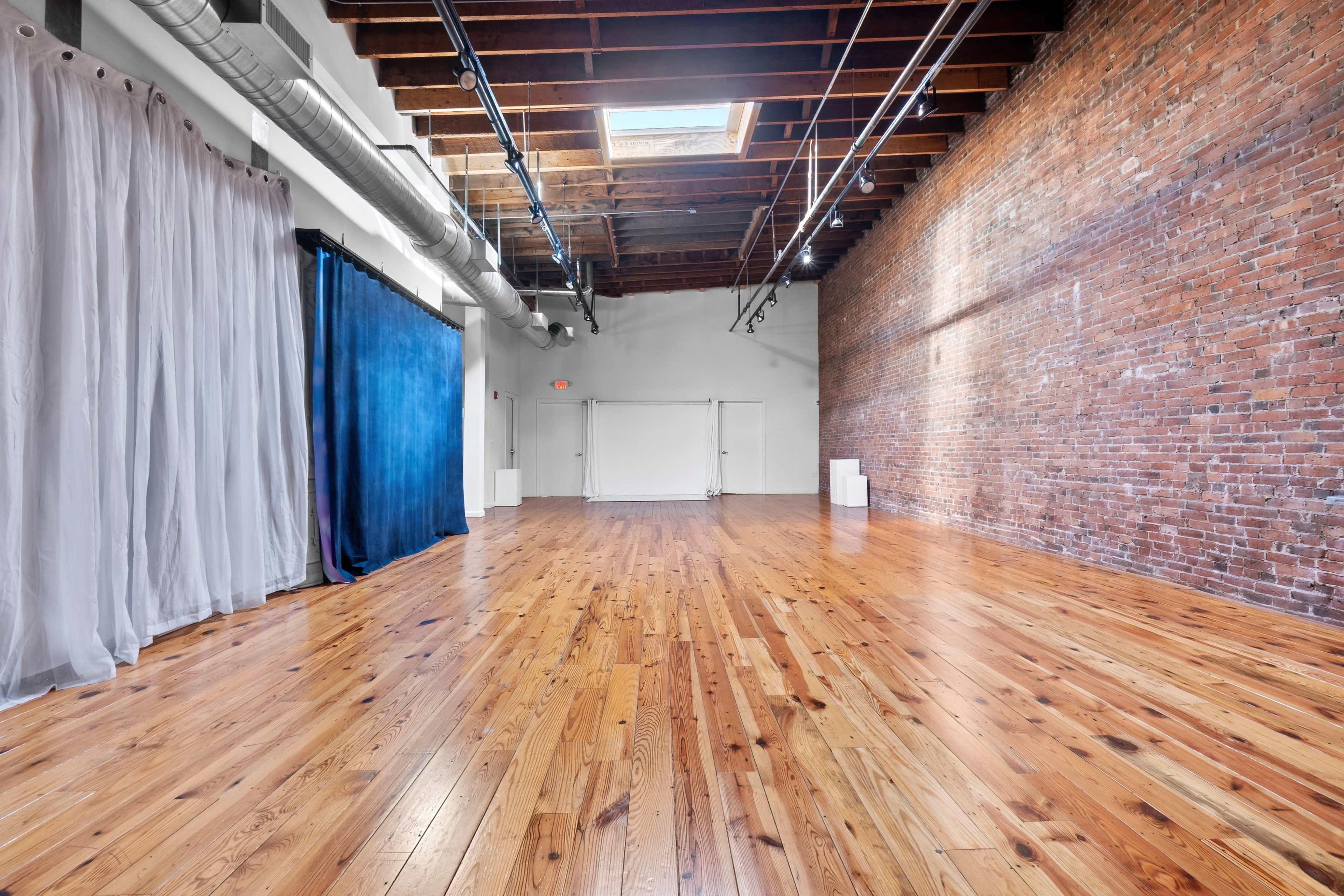 The image shows a spacious interior room with exposed brick walls, wooden floors, and large windows allowing natural light.