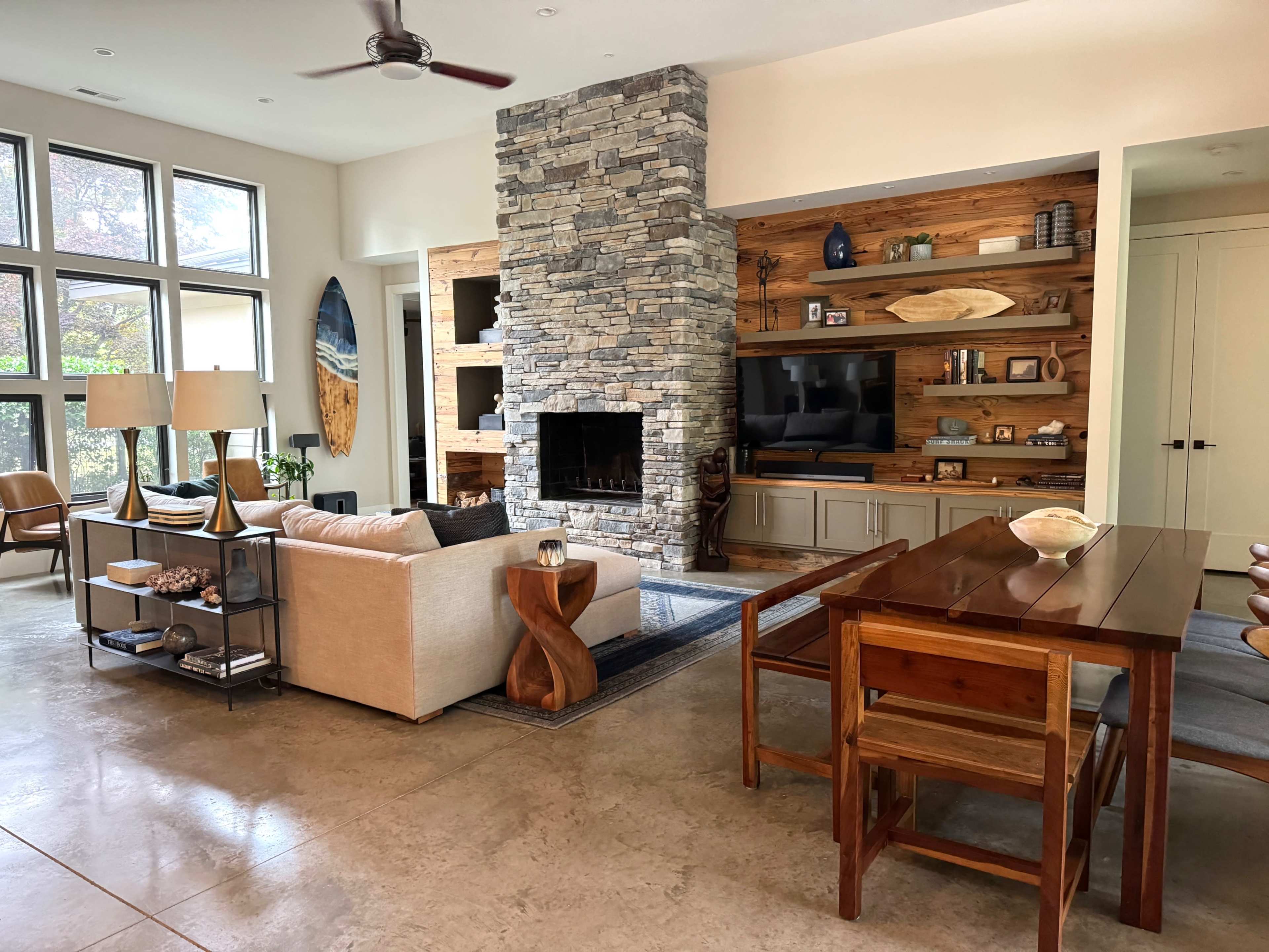 A modern living space featuring a stone fireplace, wooden shelving with decor, a beige sectional sofa, a dining table, and large windows allowing natural light.