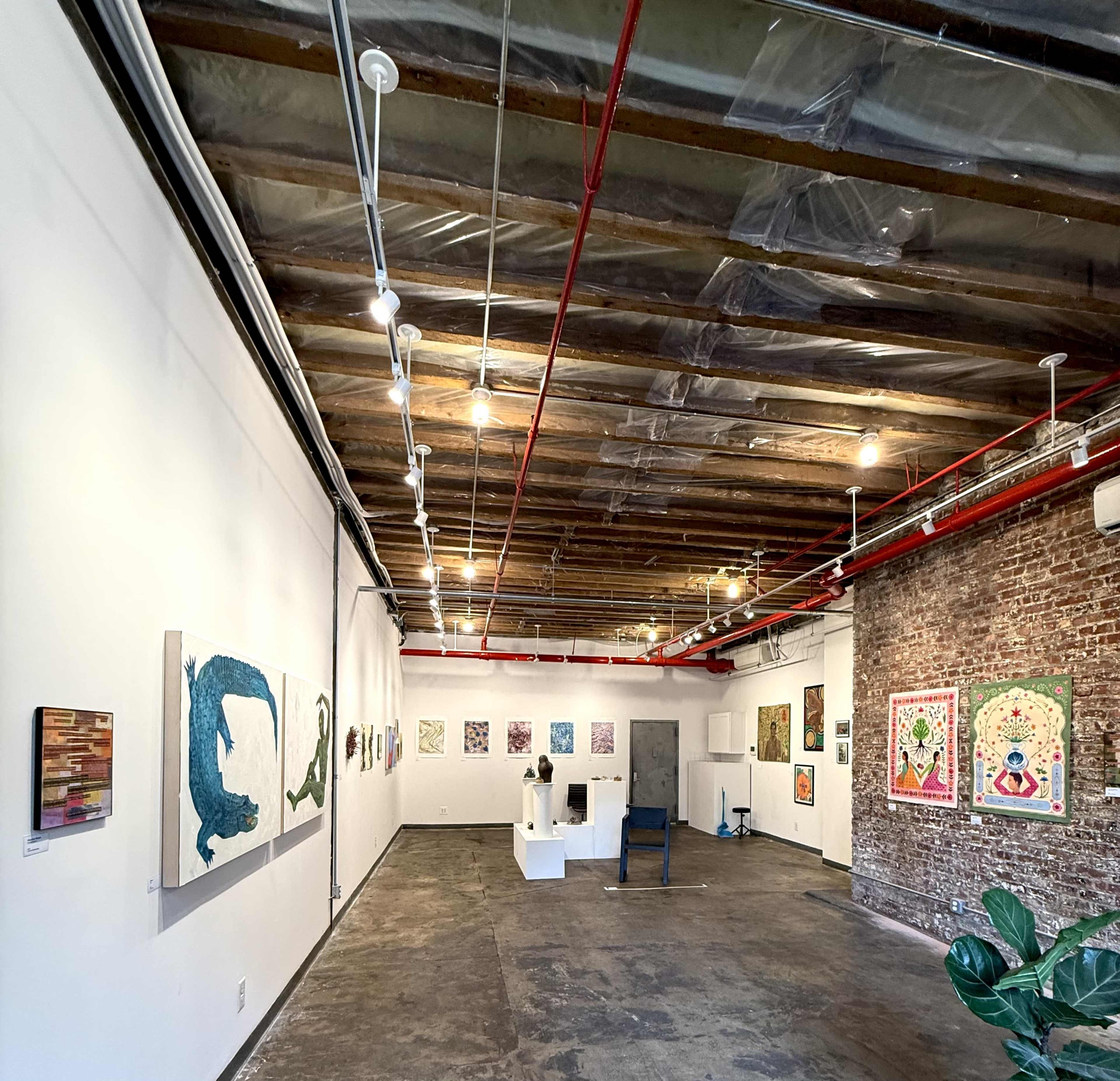 The image shows an art gallery with exposed brick walls, a polished concrete floor, and various artworks displayed on the walls.