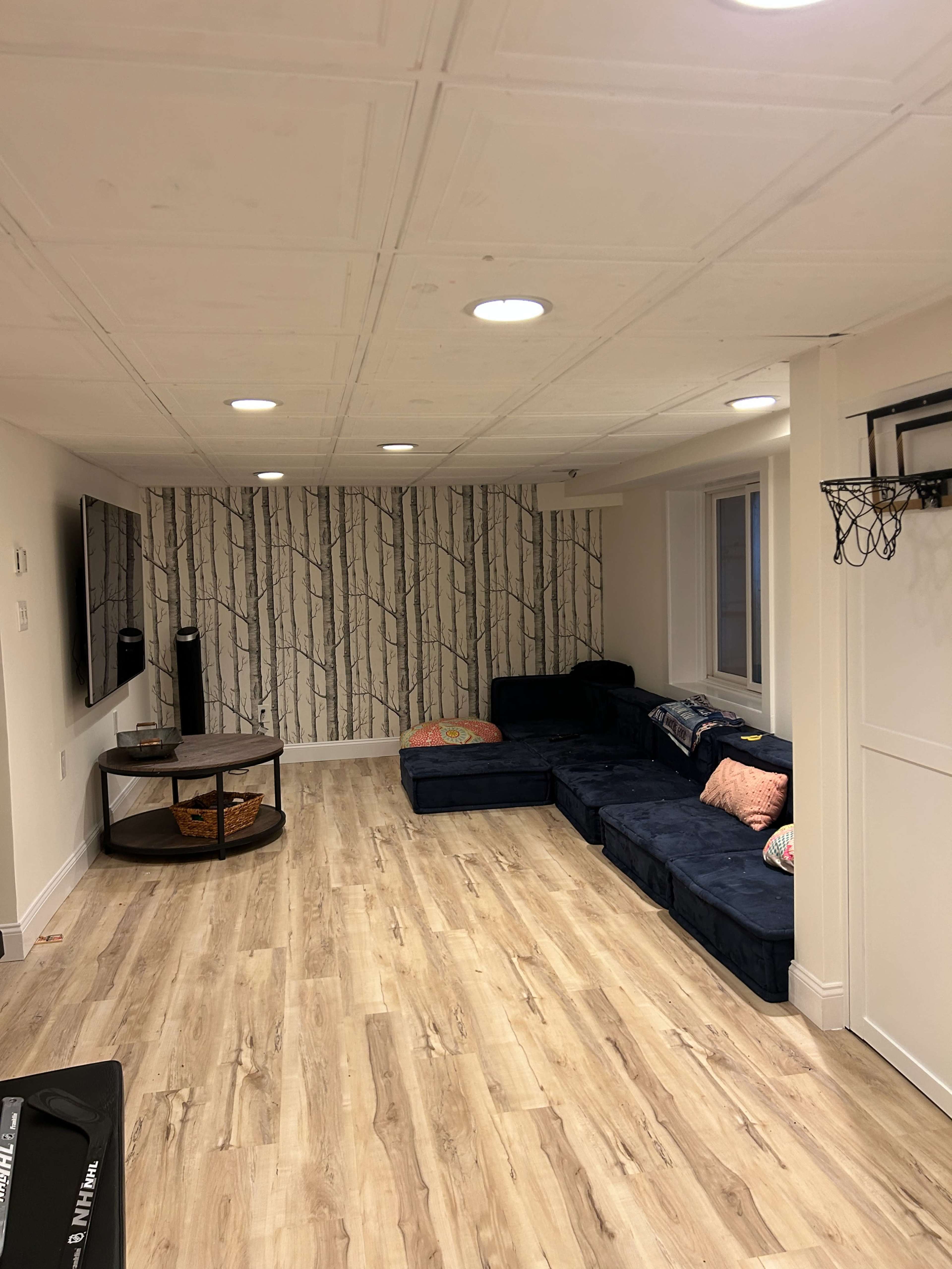 The image shows a finished basement with a sectional couch, a round coffee table, and wooden flooring, featuring a wall with a tree-patterned wallpaper.