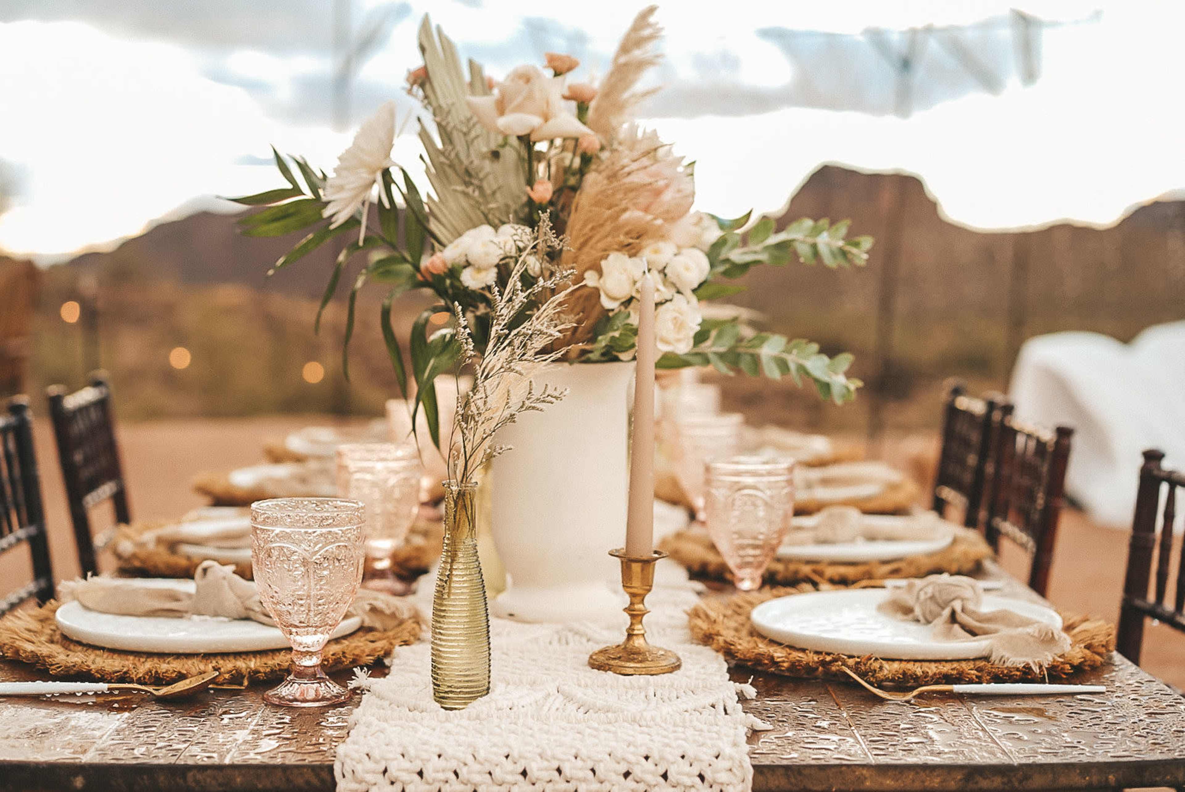 A dining table is set outdoors with elegant dishes, decorative glassware, and a floral centerpiece surrounded by mountains.