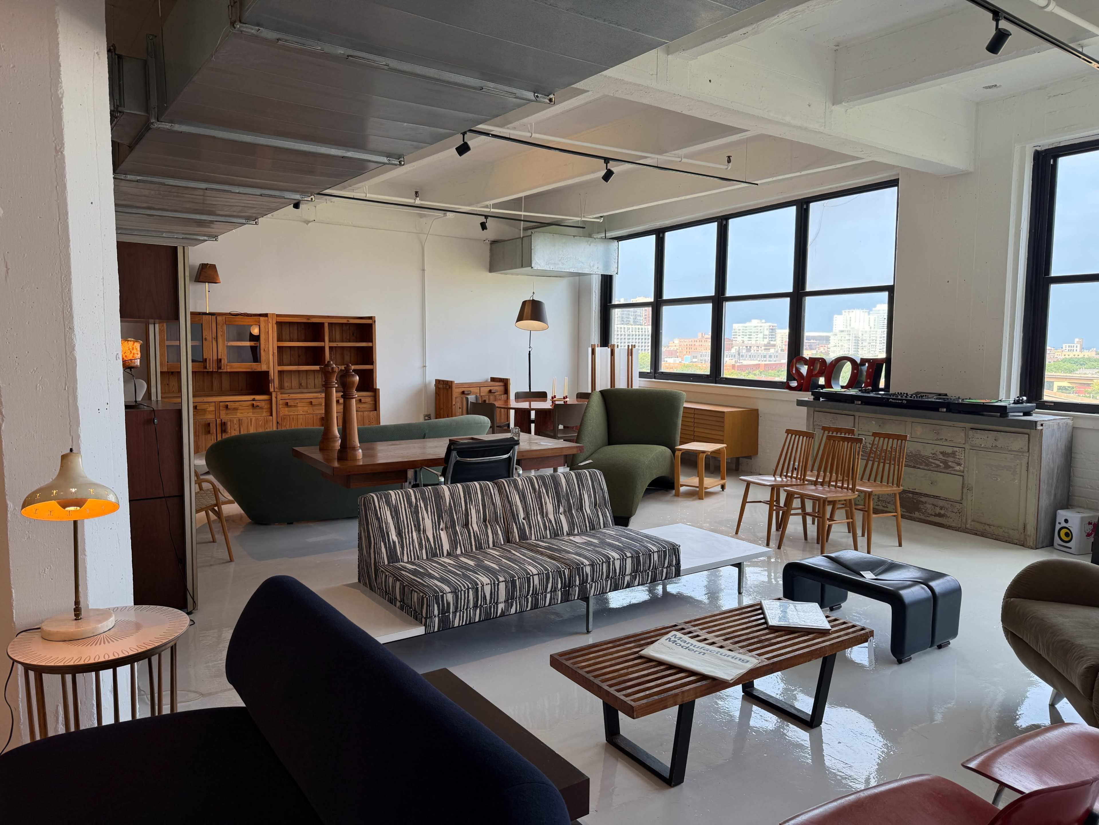 The image shows a spacious, modern interior featuring various furniture pieces, including sofas, chairs, and a dining table, with large windows allowing natural light to fill the room.