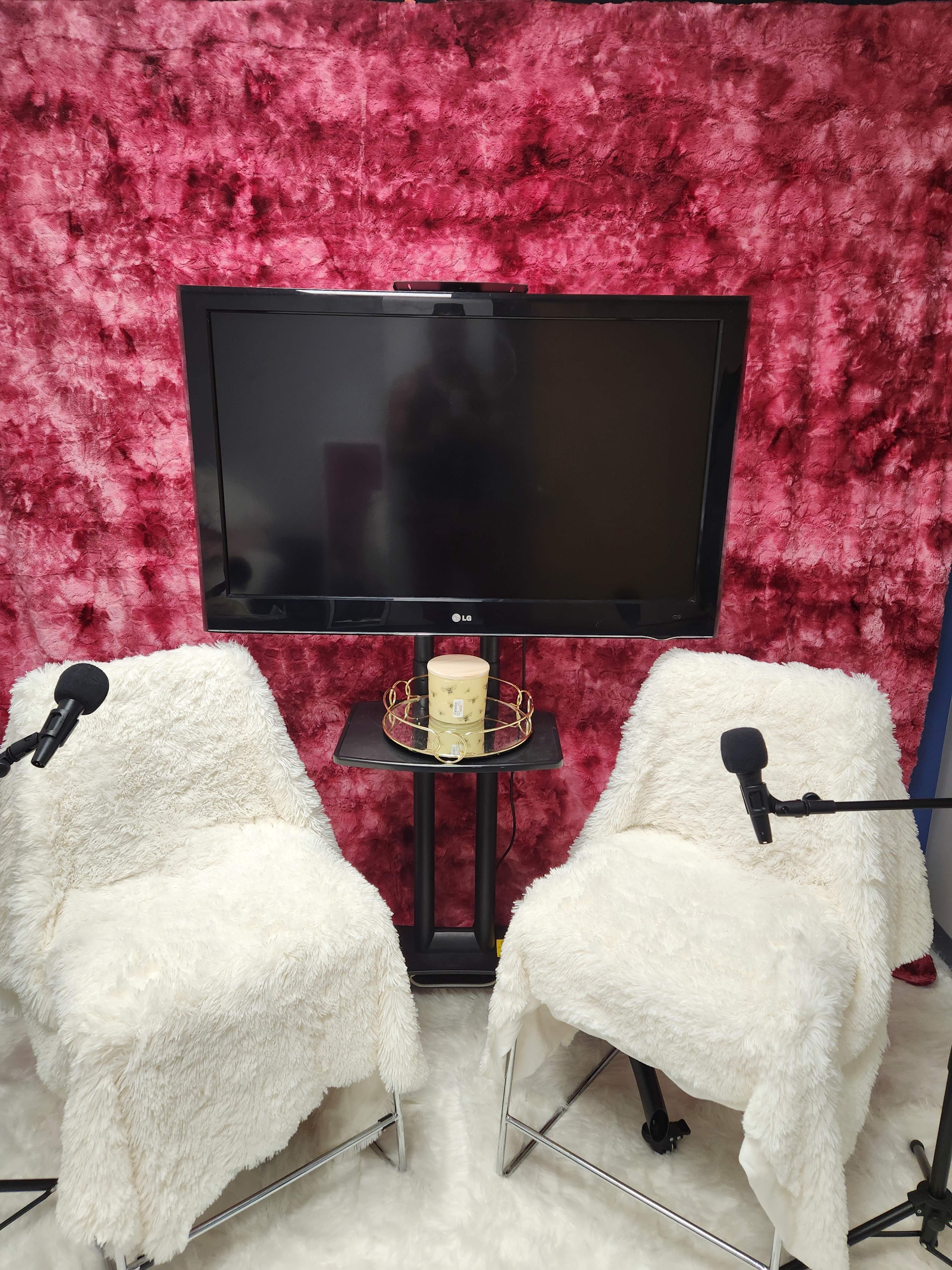 The image shows two white, furry chairs positioned facing a black television set on a stand against a pink textured backdrop.