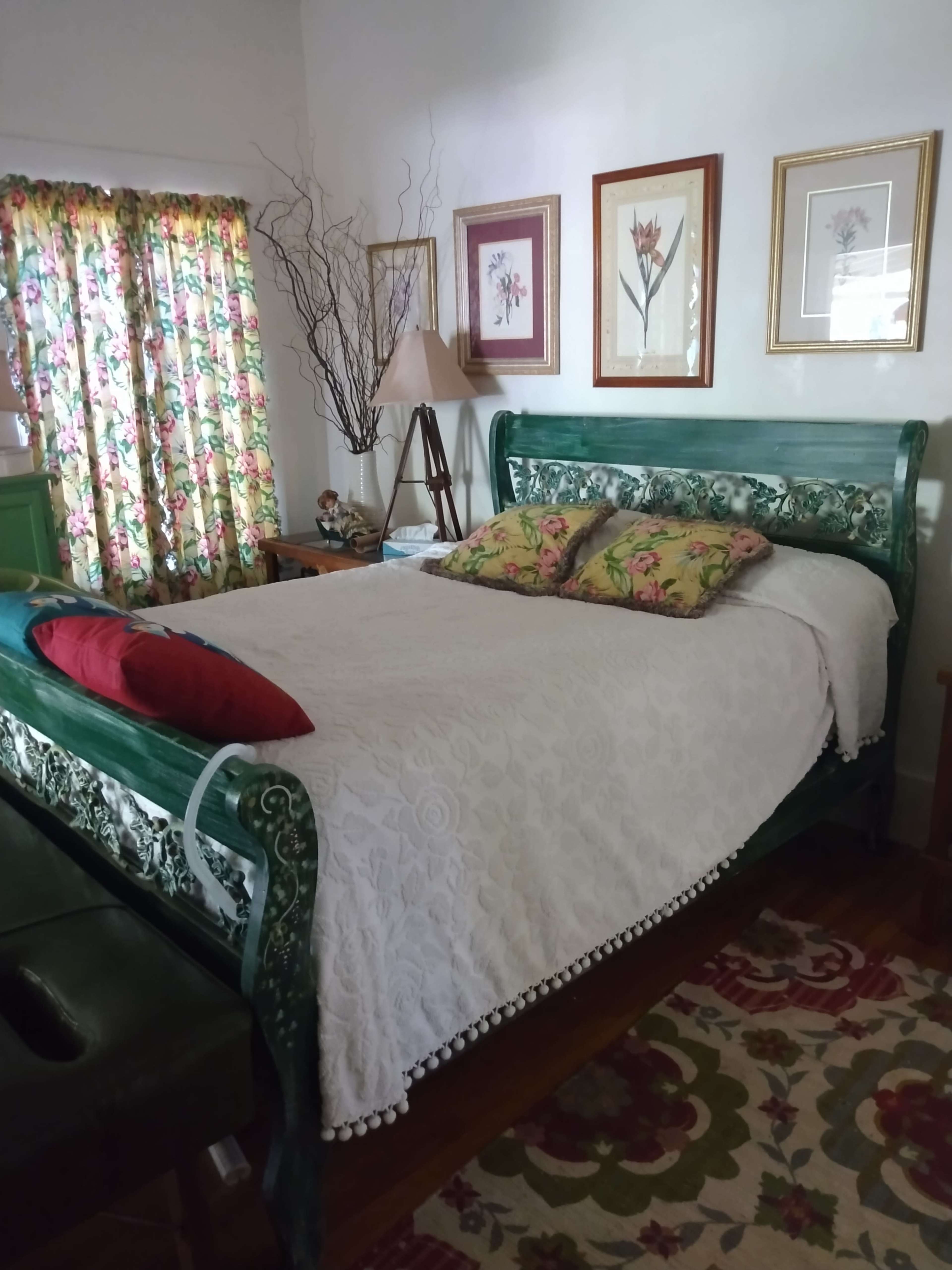 A bedroom featuring a green wooden sleigh bed with a white bedspread and decorative pillows, complemented by floral curtains and framed art on the walls.