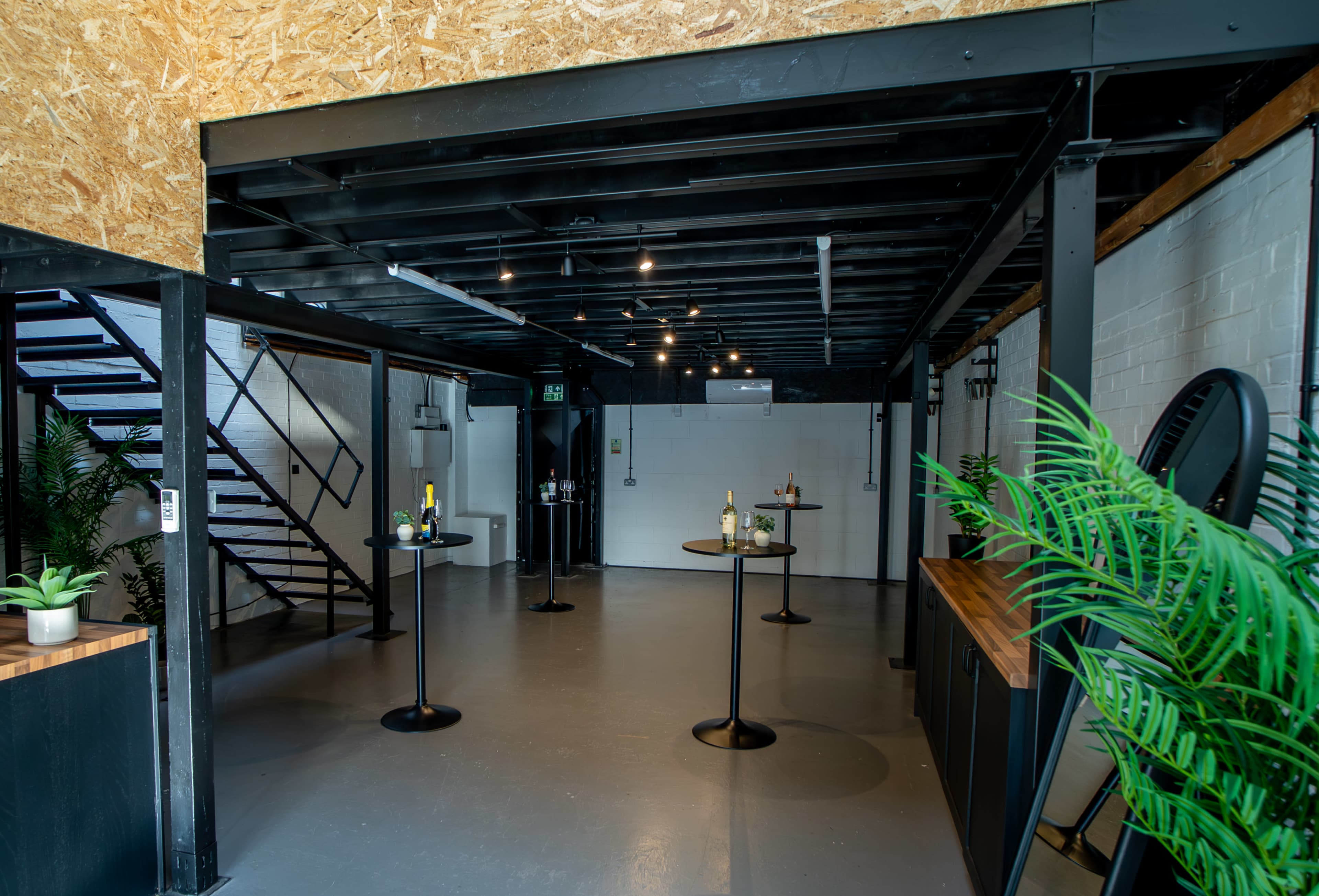 The image shows a spacious, modern indoor area with black metal beams, wooden accents, and several small standing tables set up for an event.