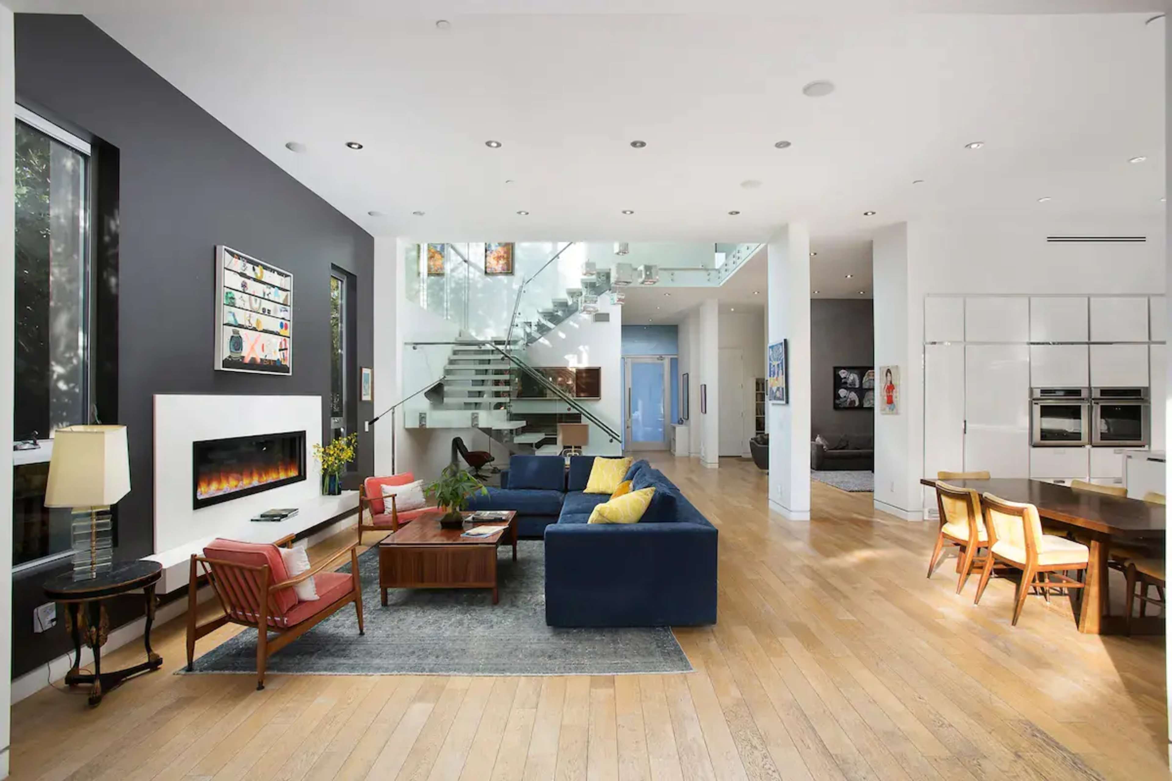 A modern open-concept living area with a sleek staircase, large windows, and a fireplace, featuring a sectional sofa, wooden dining table, and various furnishings.