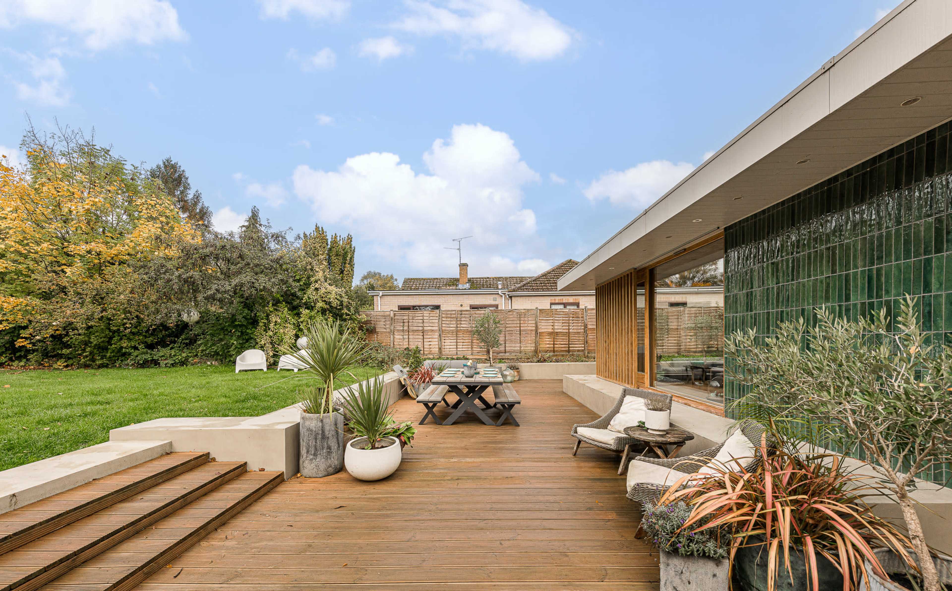 The image shows a modern backyard with a wooden deck, a seating area, and lush greenery in the background.