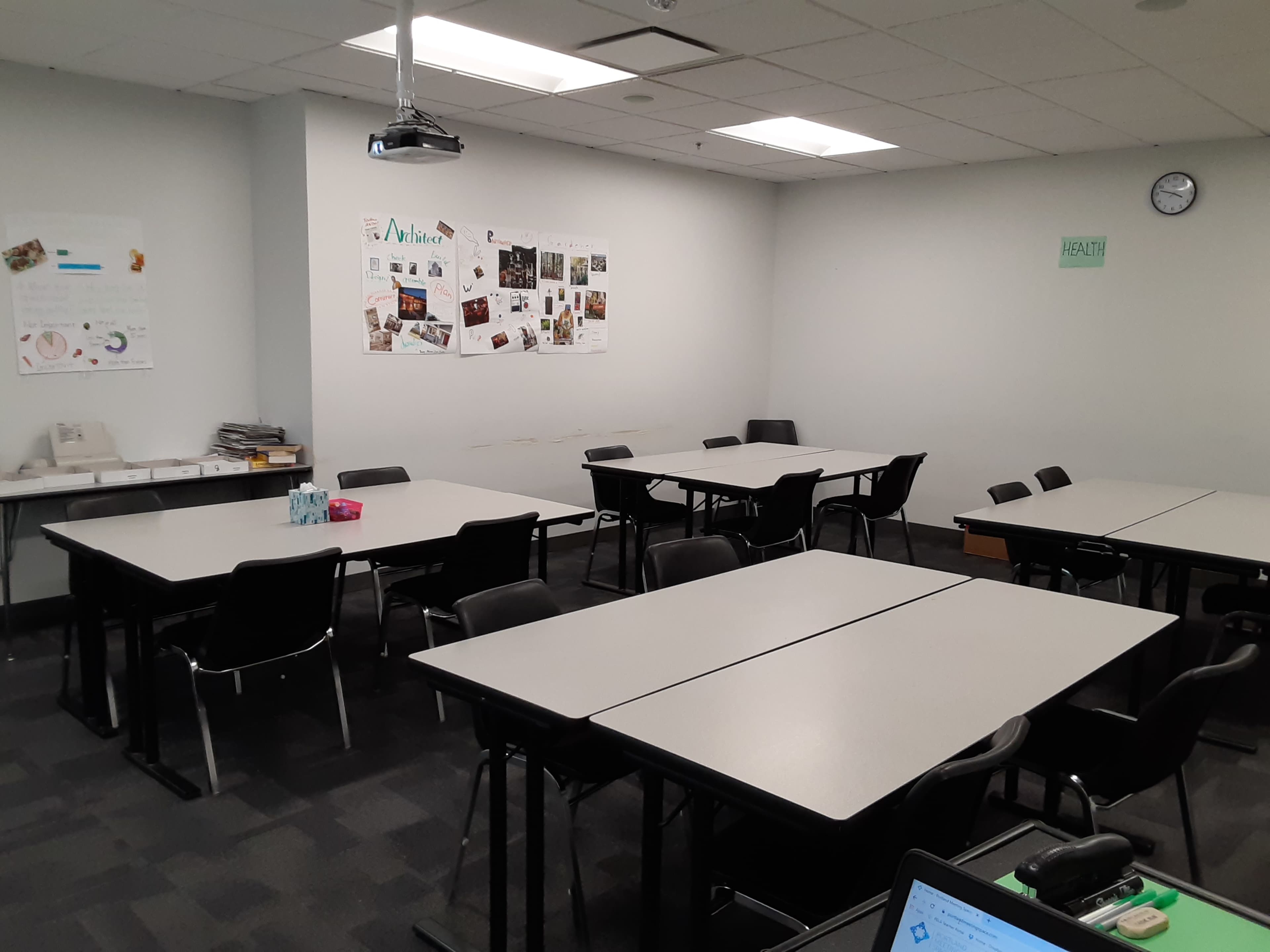 A classroom with multiple light-colored tables and black chairs arranged in an open layout, featuring posters on the wall and a clock mounted above.