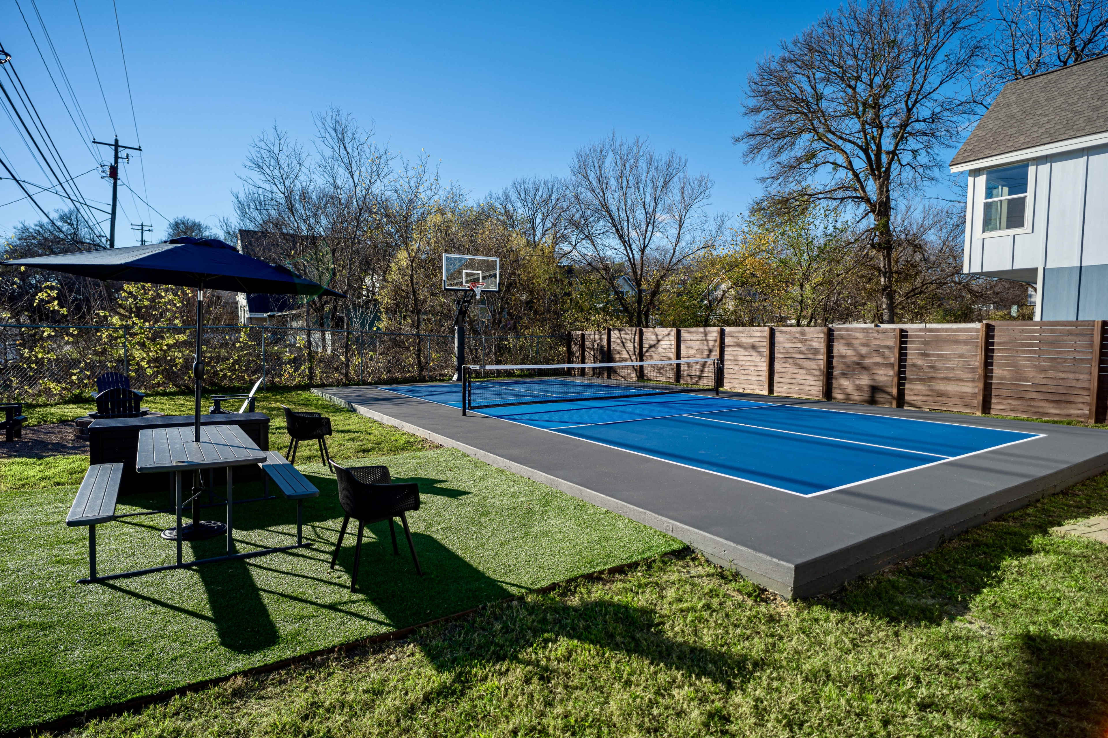 A blue basketball court is surrounded by green grass and a seating area with a picnic table and chairs under an umbrella.