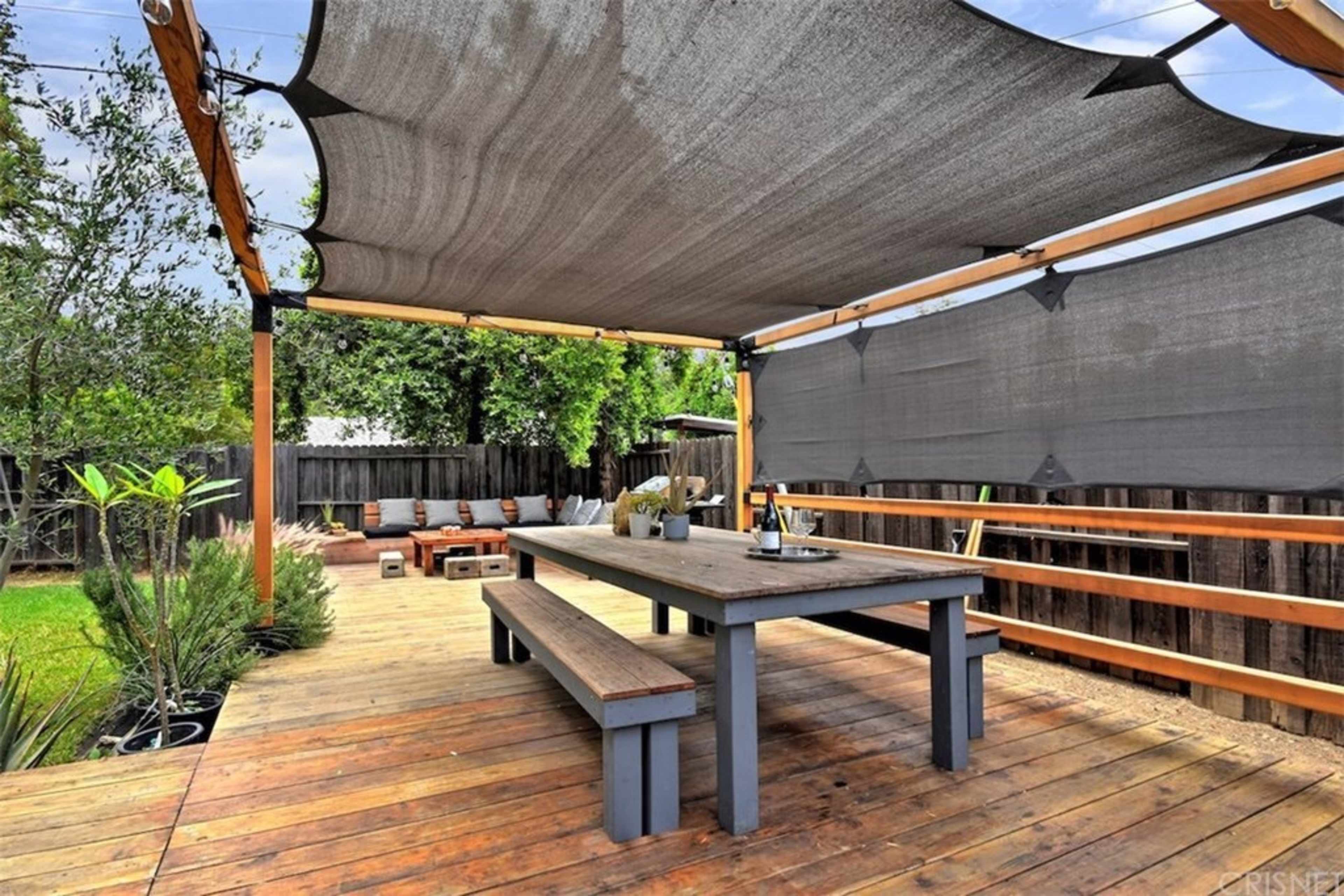 The image shows a wooden deck with a large table and a shaded area covered by a sail-like awning, surrounded by greenery and seating.
