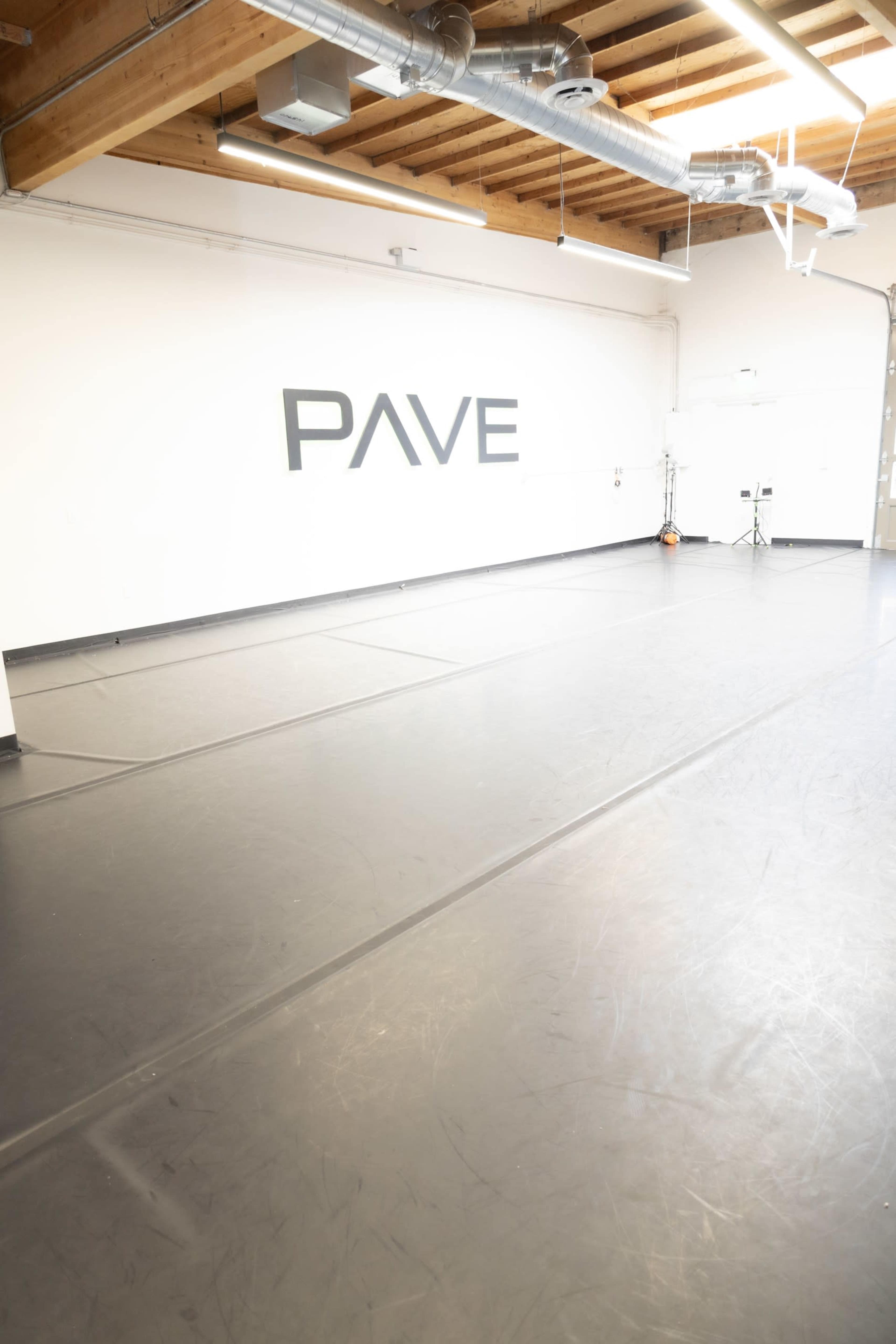 The image shows an empty studio space with a black floor and the word "PAVE" displayed on the white wall.