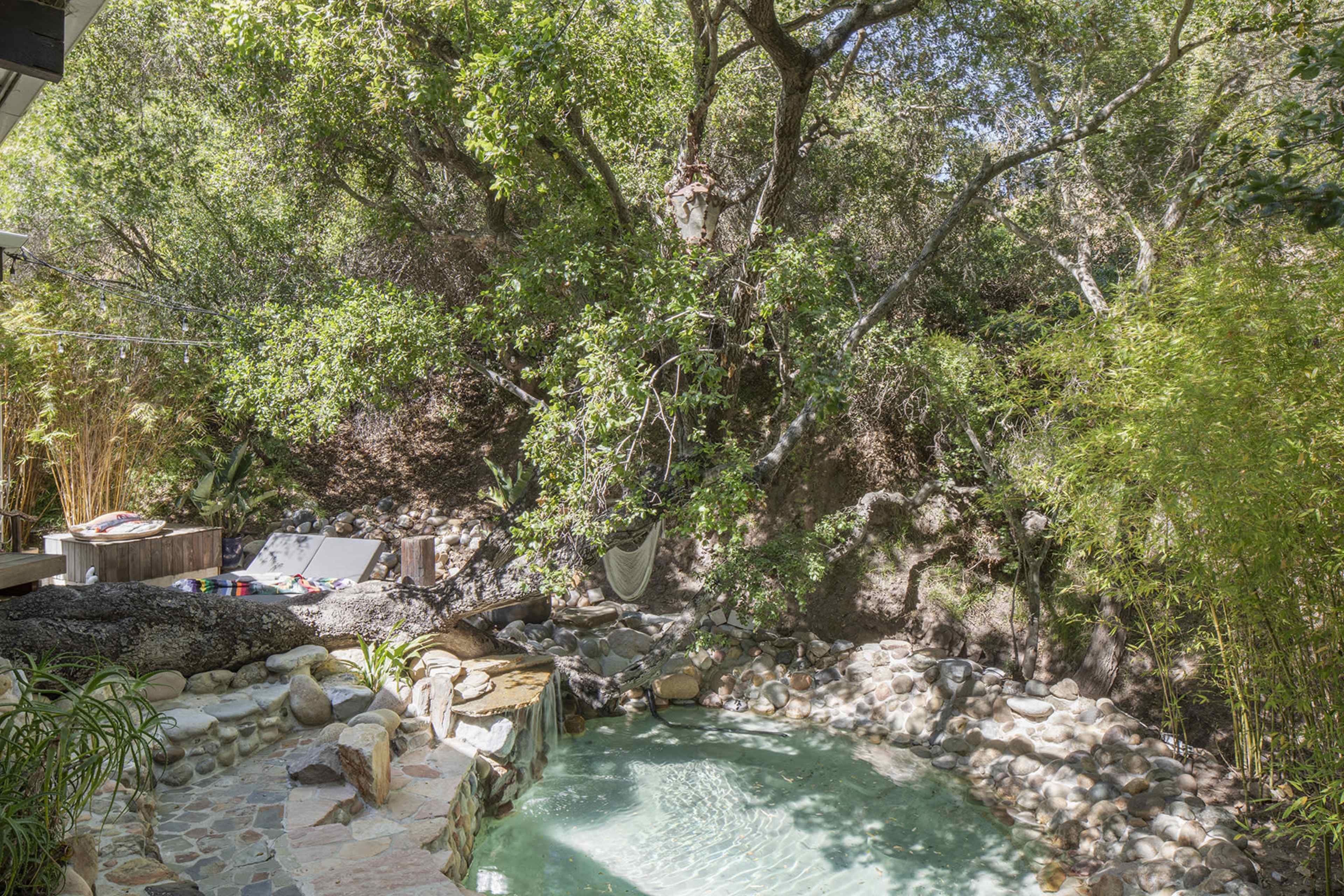 A small, natural swimming pool surrounded by rocks and lush greenery, with lounge chairs and a hammock nearby.