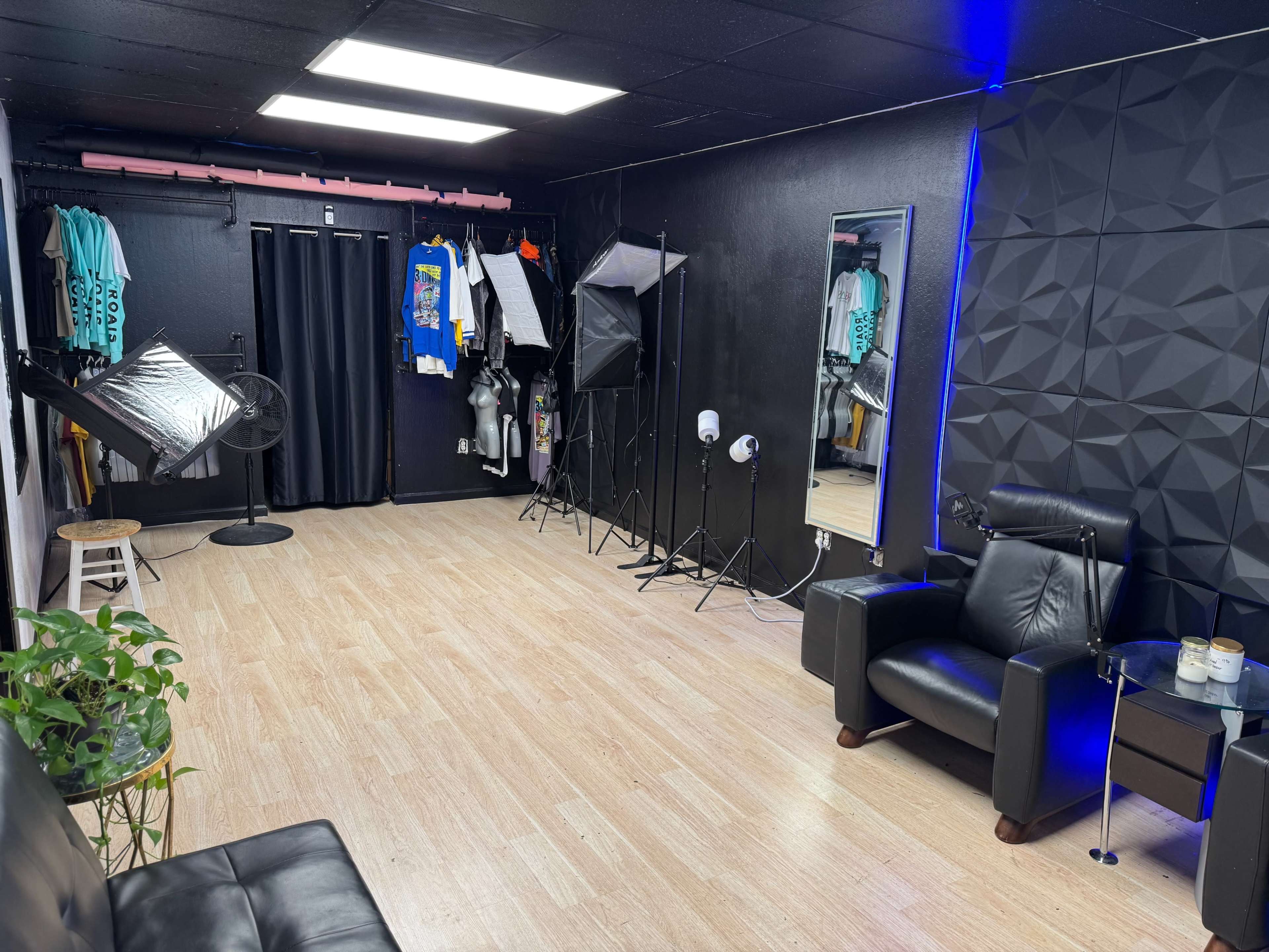 The image shows a photography studio with a black wall, professional lighting equipment, clothing on display, and a seating area with a plant.