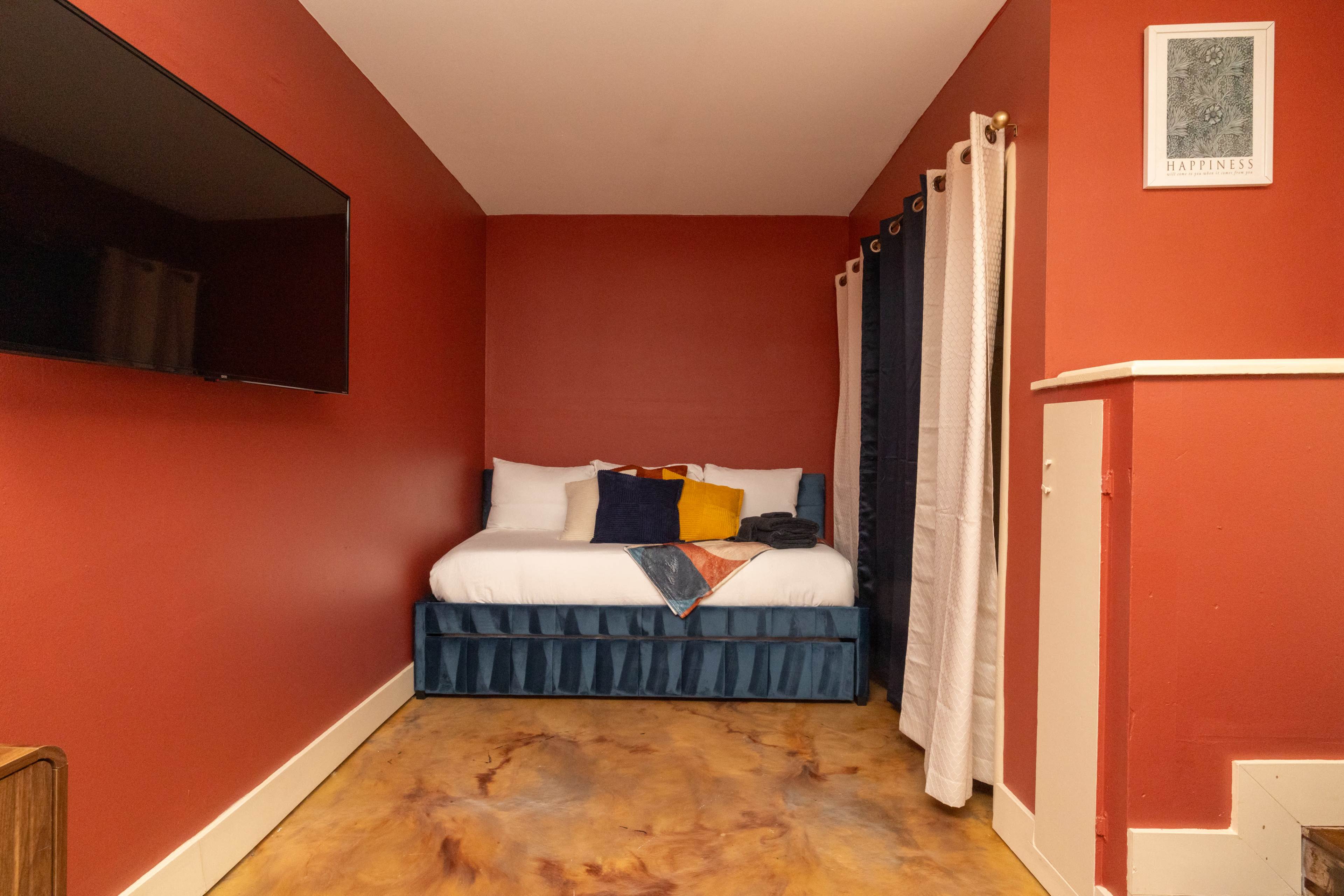 The image shows a small bedroom with a red wall, a bed adorned with pillows, and a TV mounted on the wall.