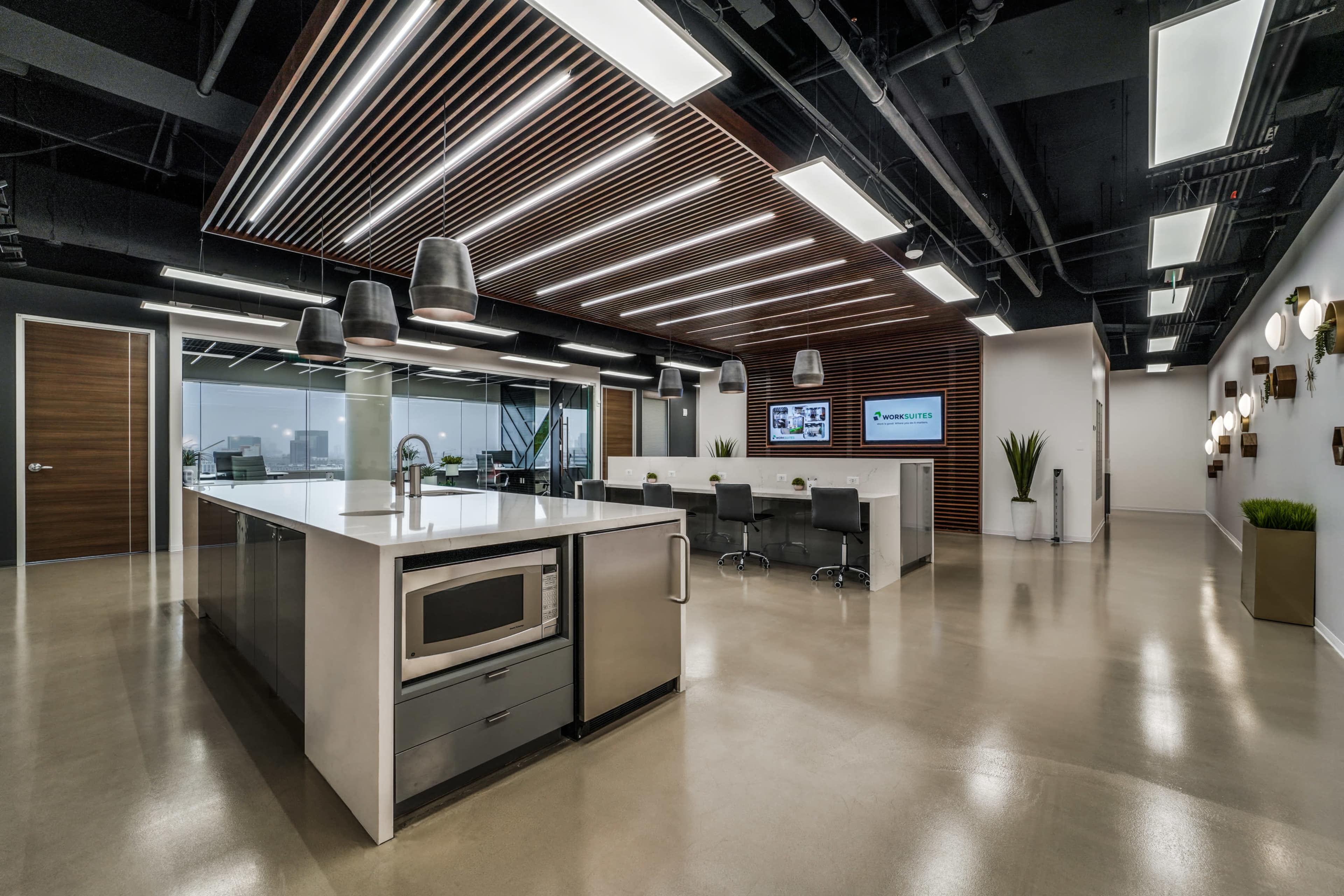 The image shows a modern office kitchen area with a large island containing a microwave and sleek cabinetry, surrounded by contemporary seating and decorative lighting.