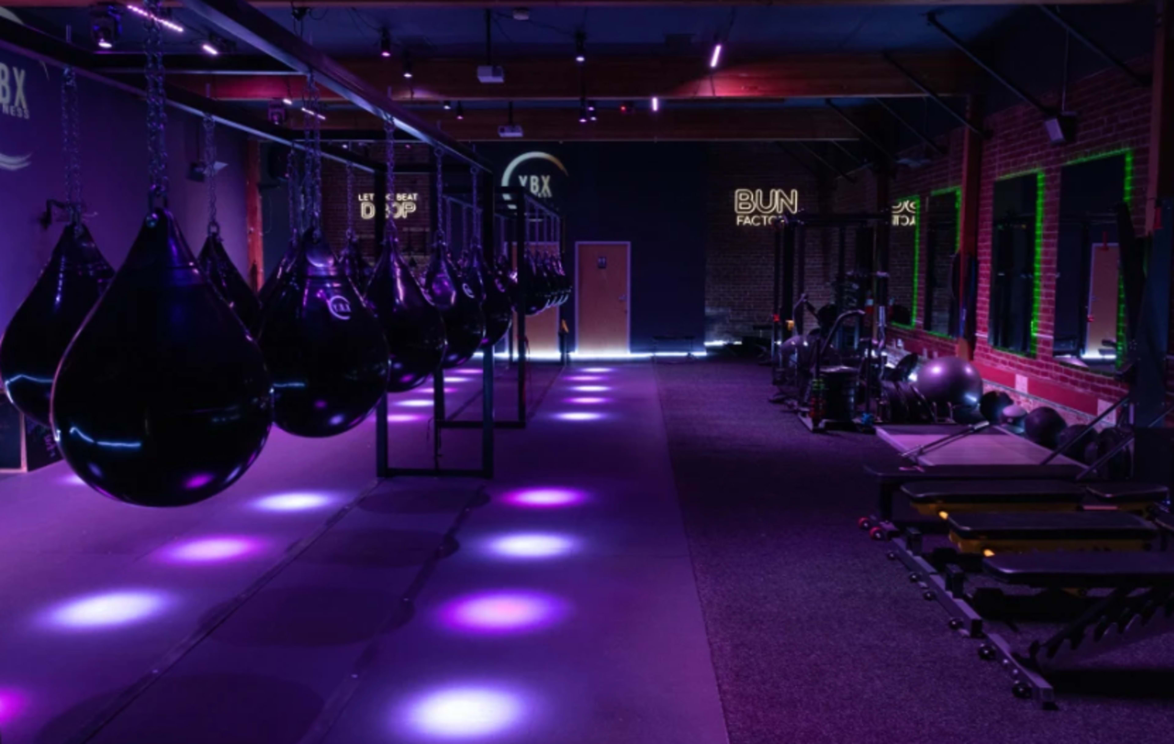 The image shows a modern fitness studio with hanging black punching bags and purple lighting illuminating the workout area.