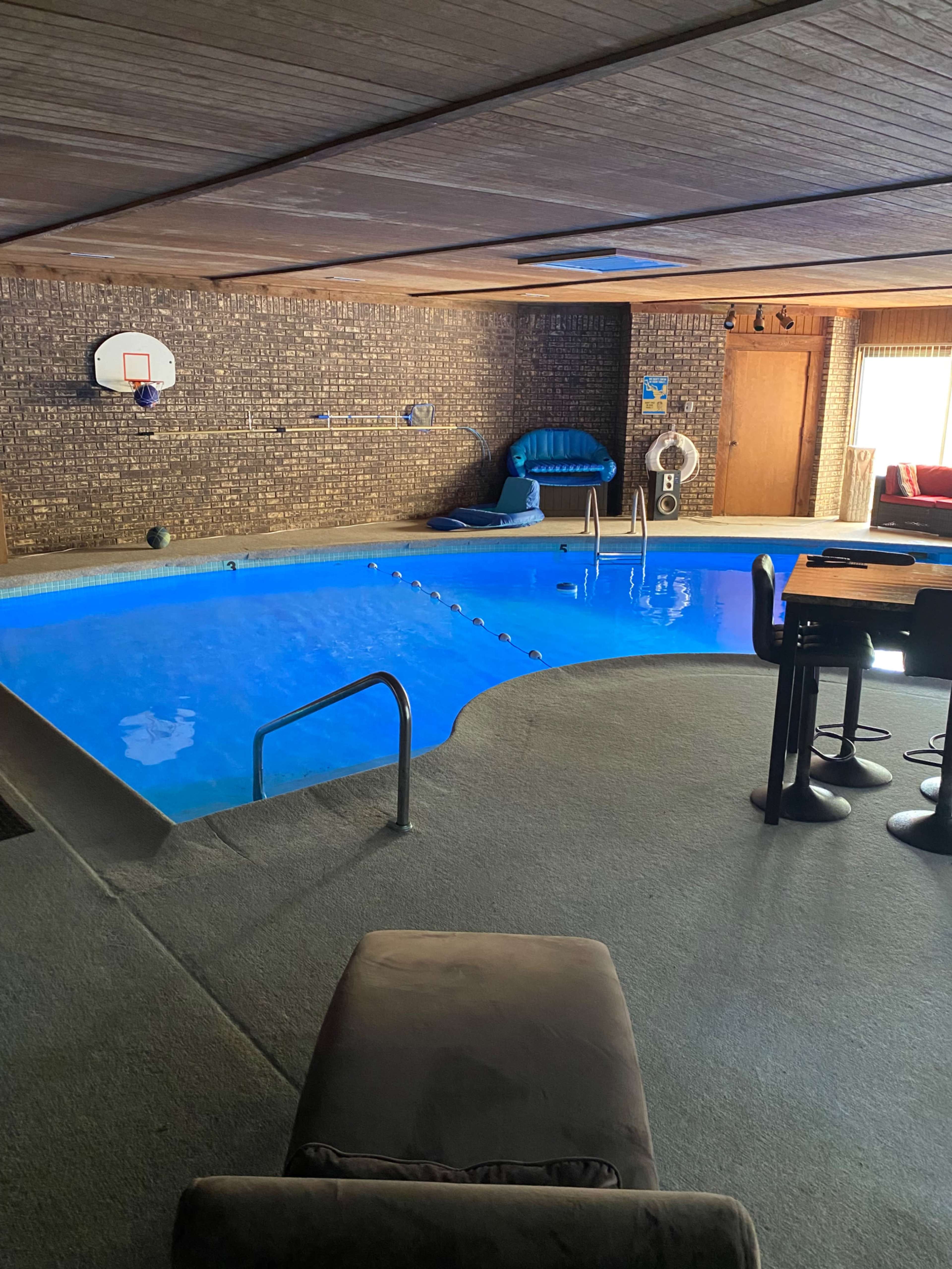 An indoor pool with a basketball hoop, surrounded by a lounge area and a bar-height table setup.