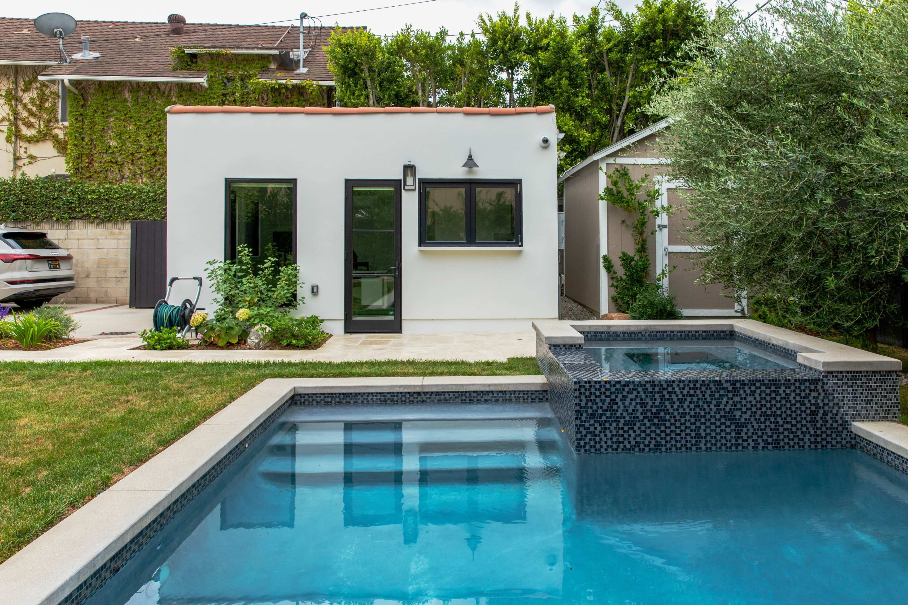 A modern backyard featuring a swimming pool, a spa, a small white building, and lush greenery.