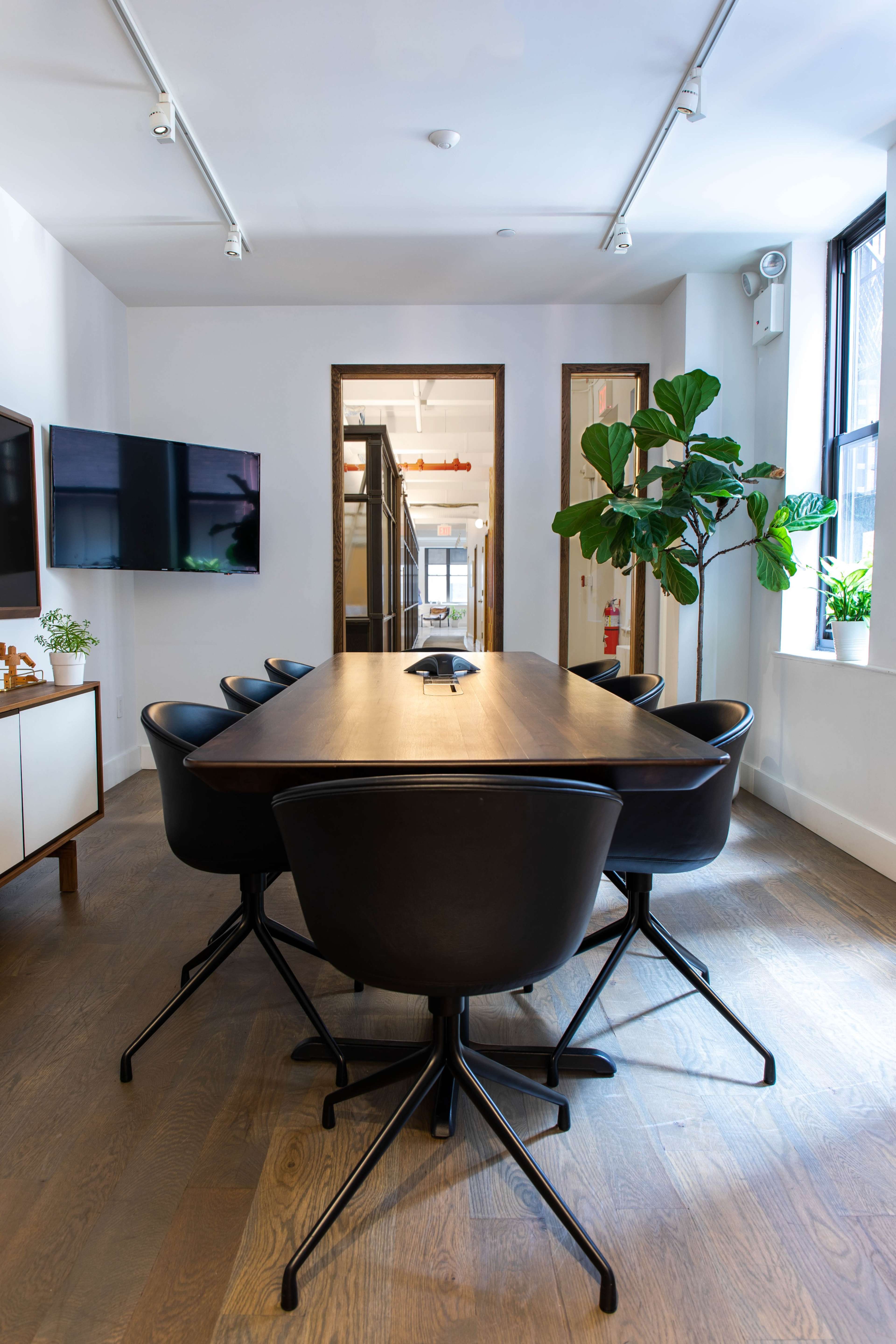 A modern conference room features a long wooden table surrounded by black chairs, with large windows and plants adding to the decor.