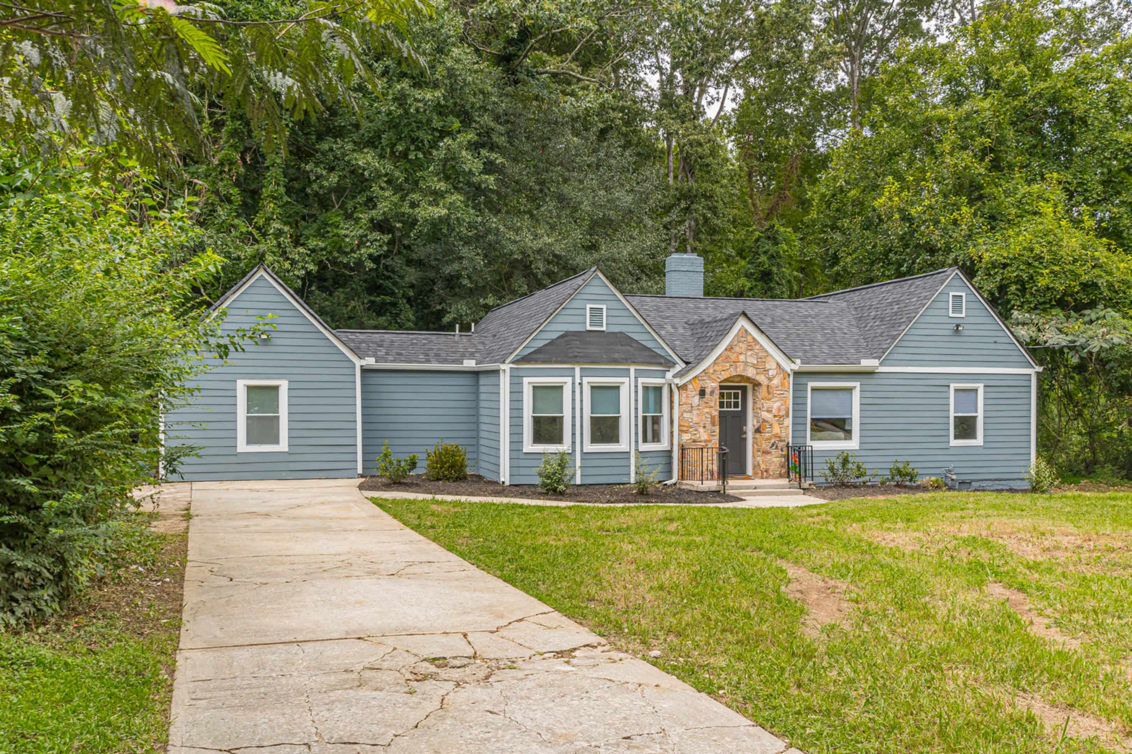 A blue, single-story house with a stone accent near the entrance, surrounded by greenery and a paved driveway.