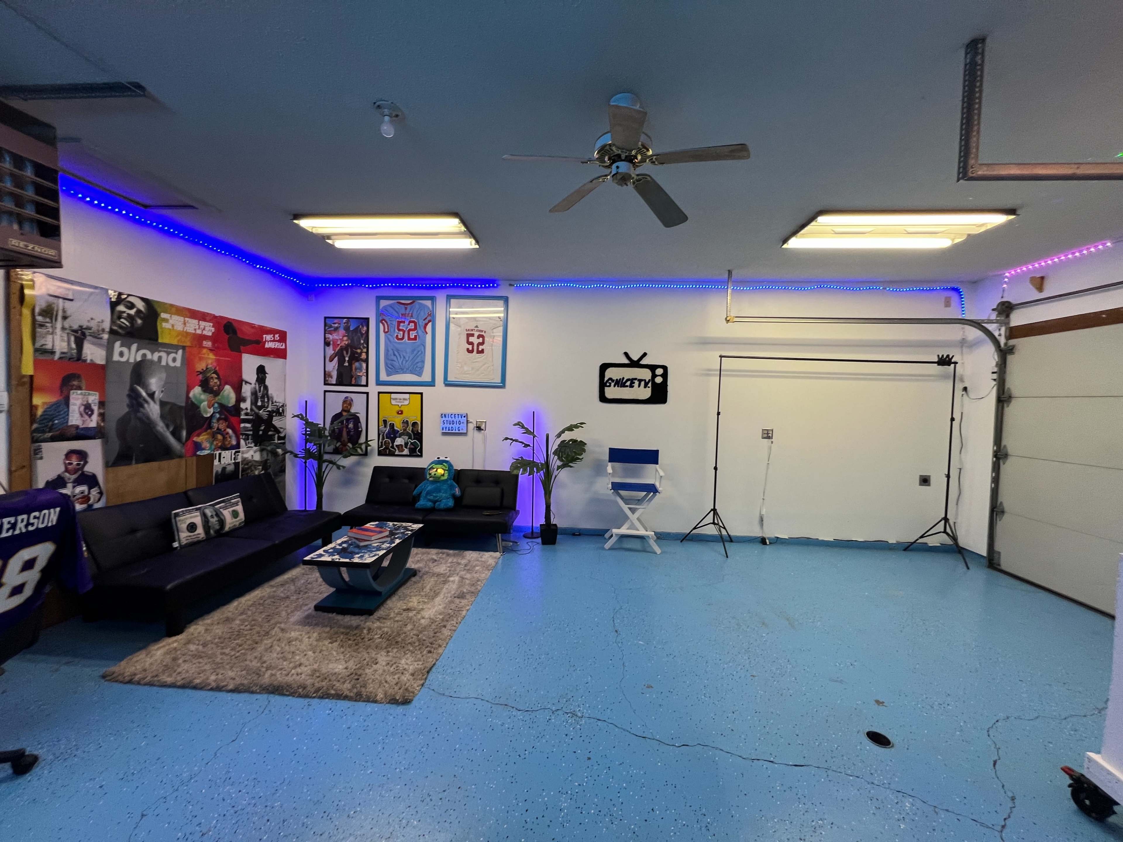 The image shows a garage interior featuring a seating area with couches, colorful wall decorations, a ceiling fan, and blue-painted flooring.