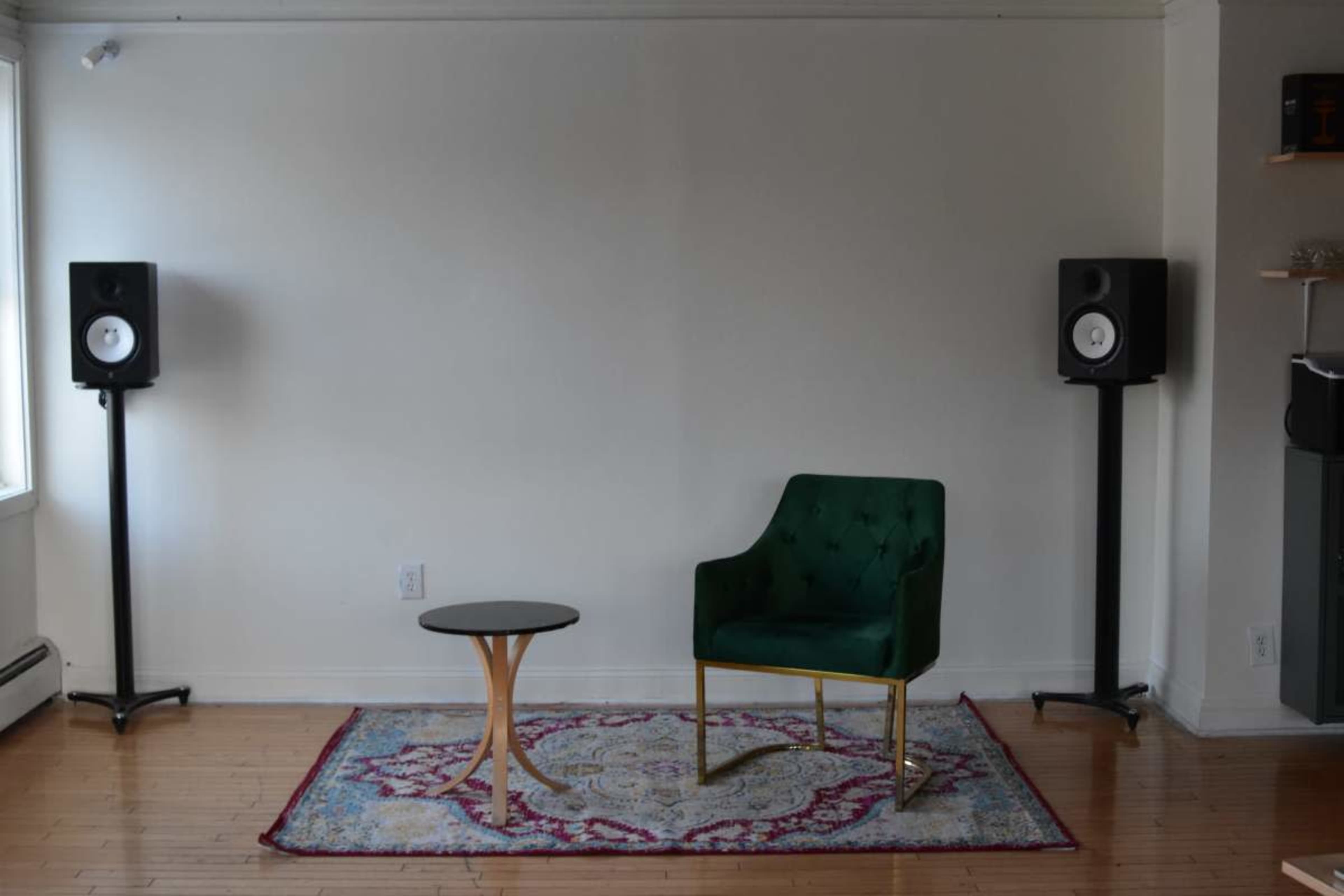 The image shows a minimalist room featuring a green chair and a small round table on a patterned rug, flanked by two speakers.