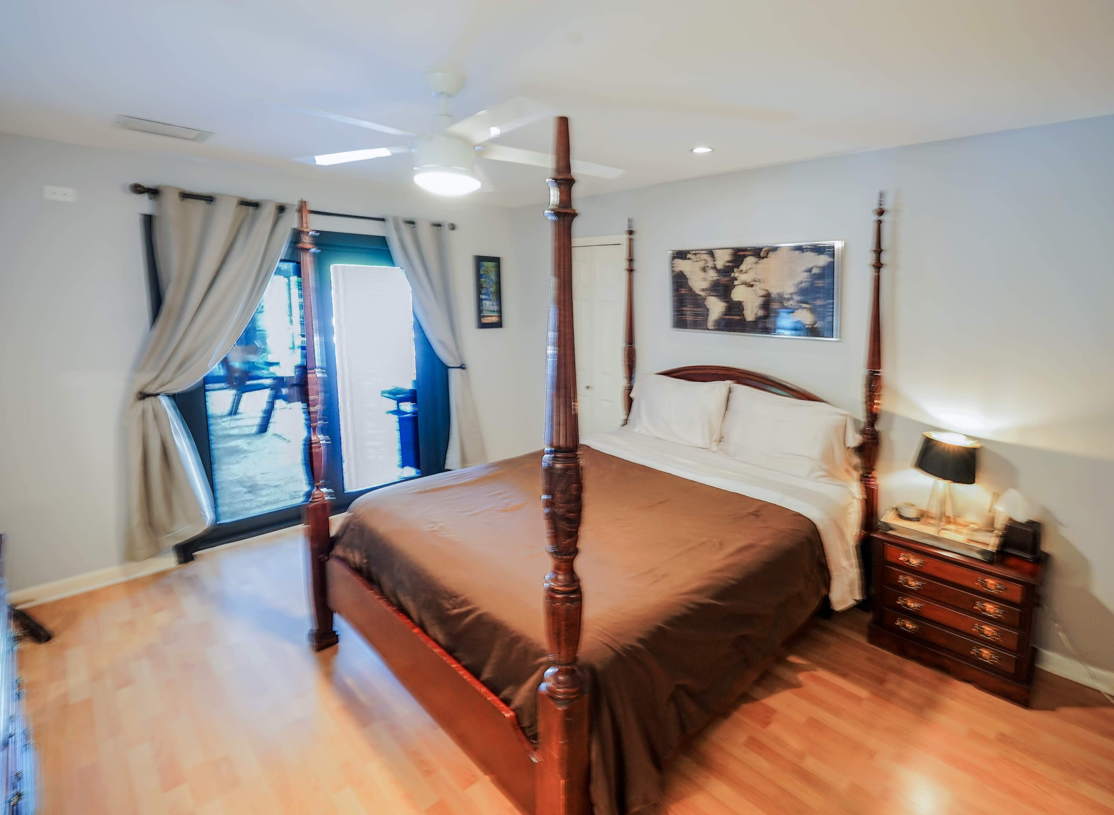 The image shows a bedroom featuring a four-poster bed, a dresser with a lamp, and large windows with curtains.
