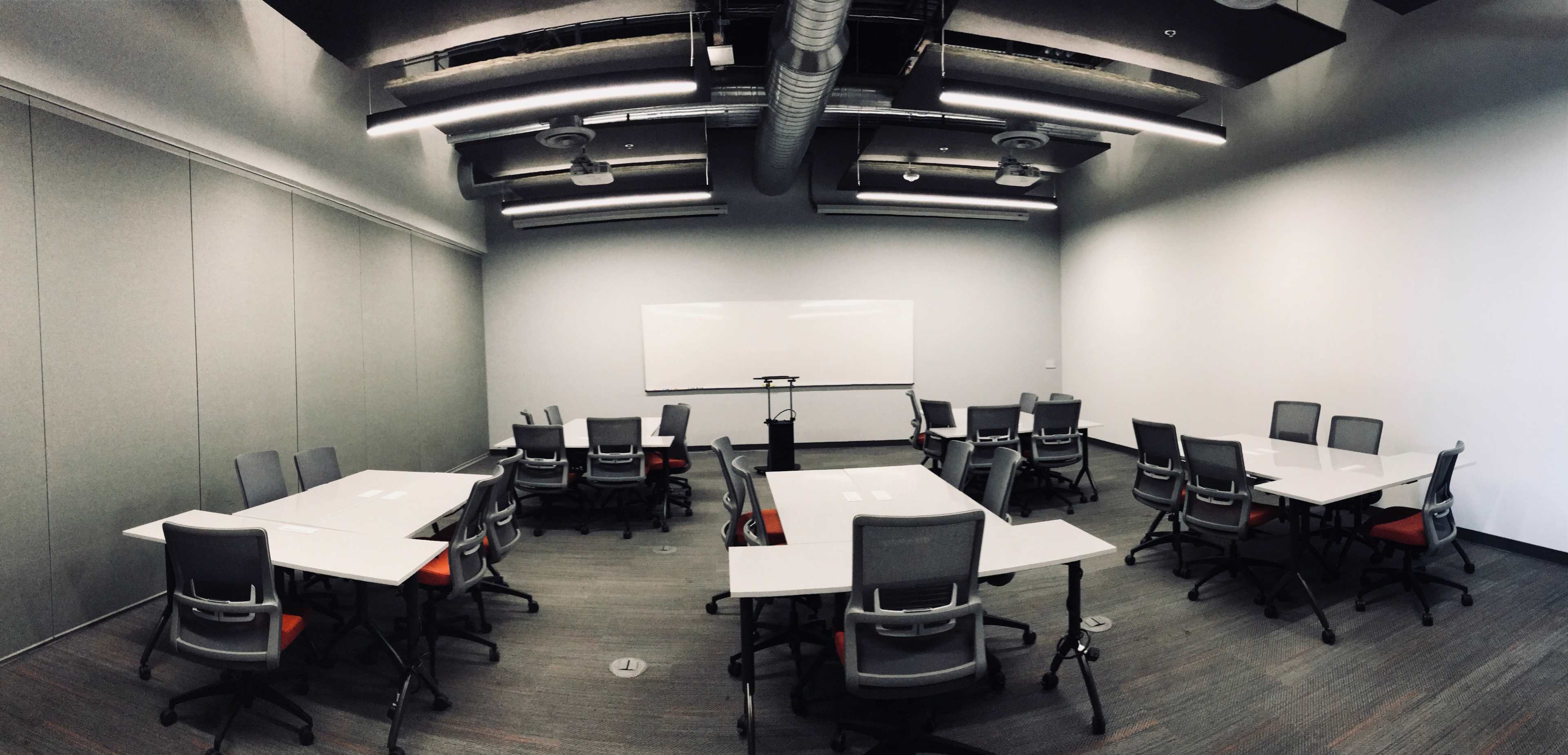 A modern classroom features multiple tables and chairs arranged in a circular layout, with a whiteboard at the front and metal ductwork visible on the ceiling.