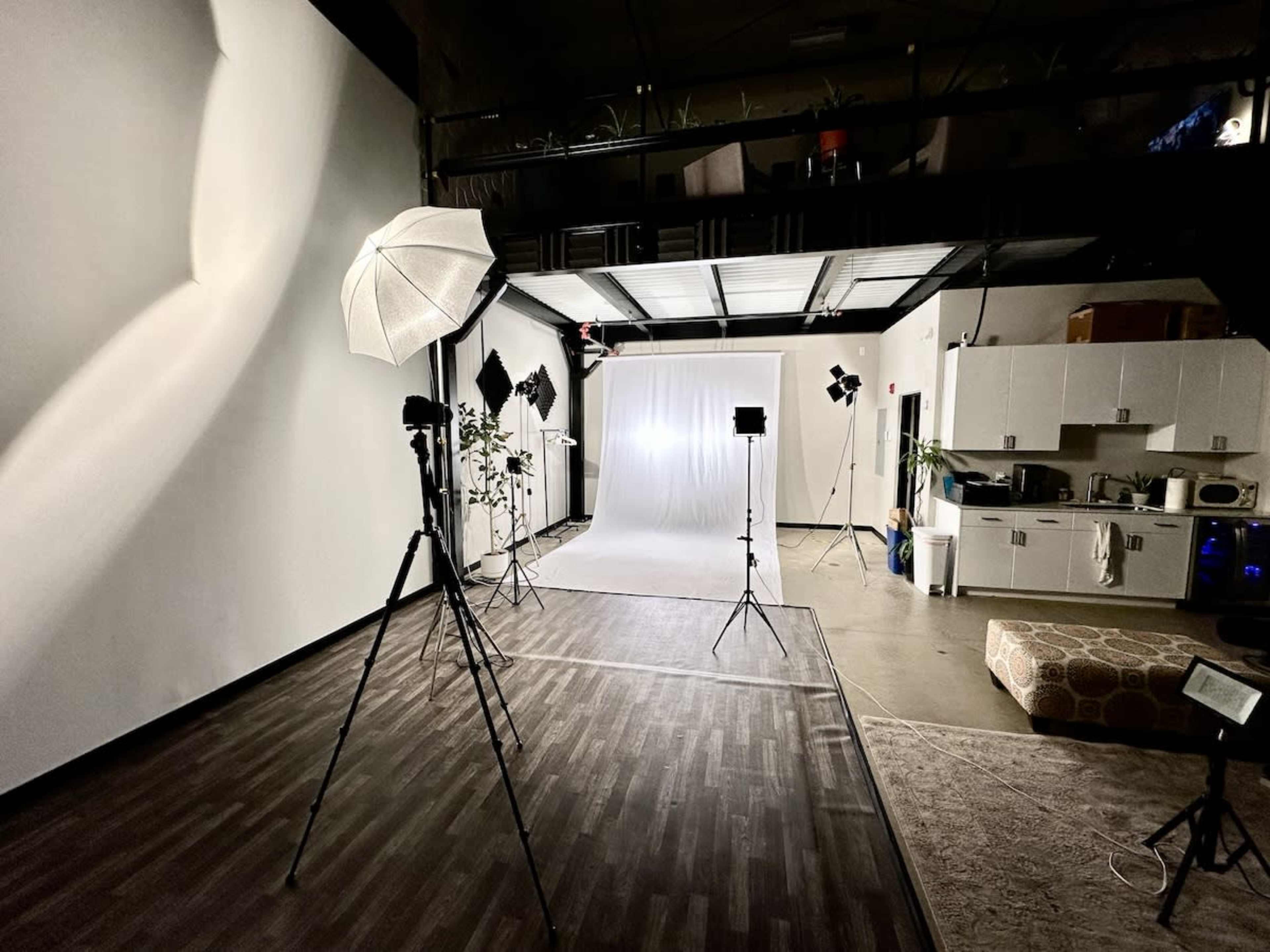 The image shows a photography studio equipped with lighting equipment, a backdrop, and an adjacent kitchenette area.