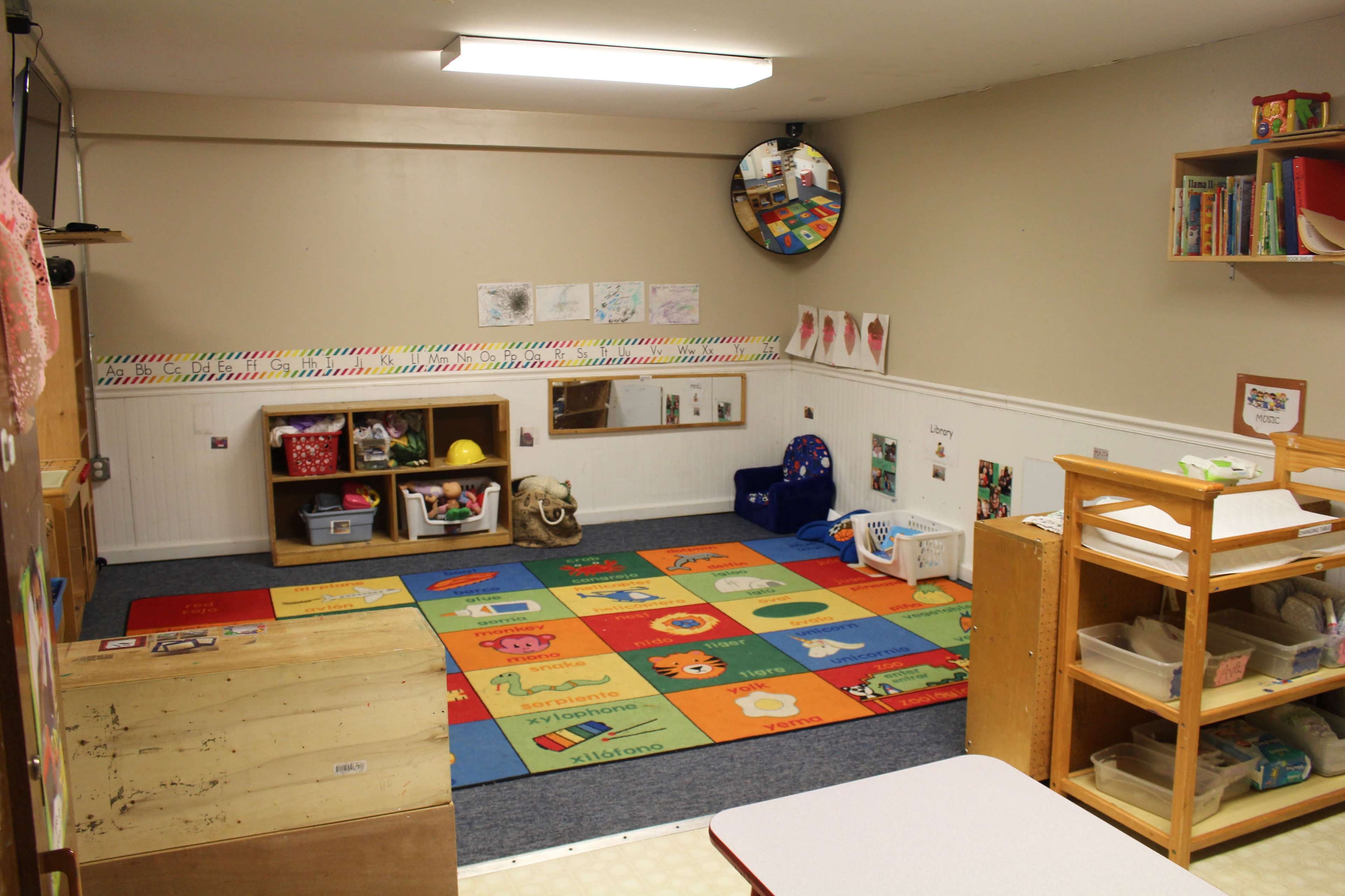 A classroom with colorful carpet tiles, shelves filled with educational materials, and a play area equipped with various toys.