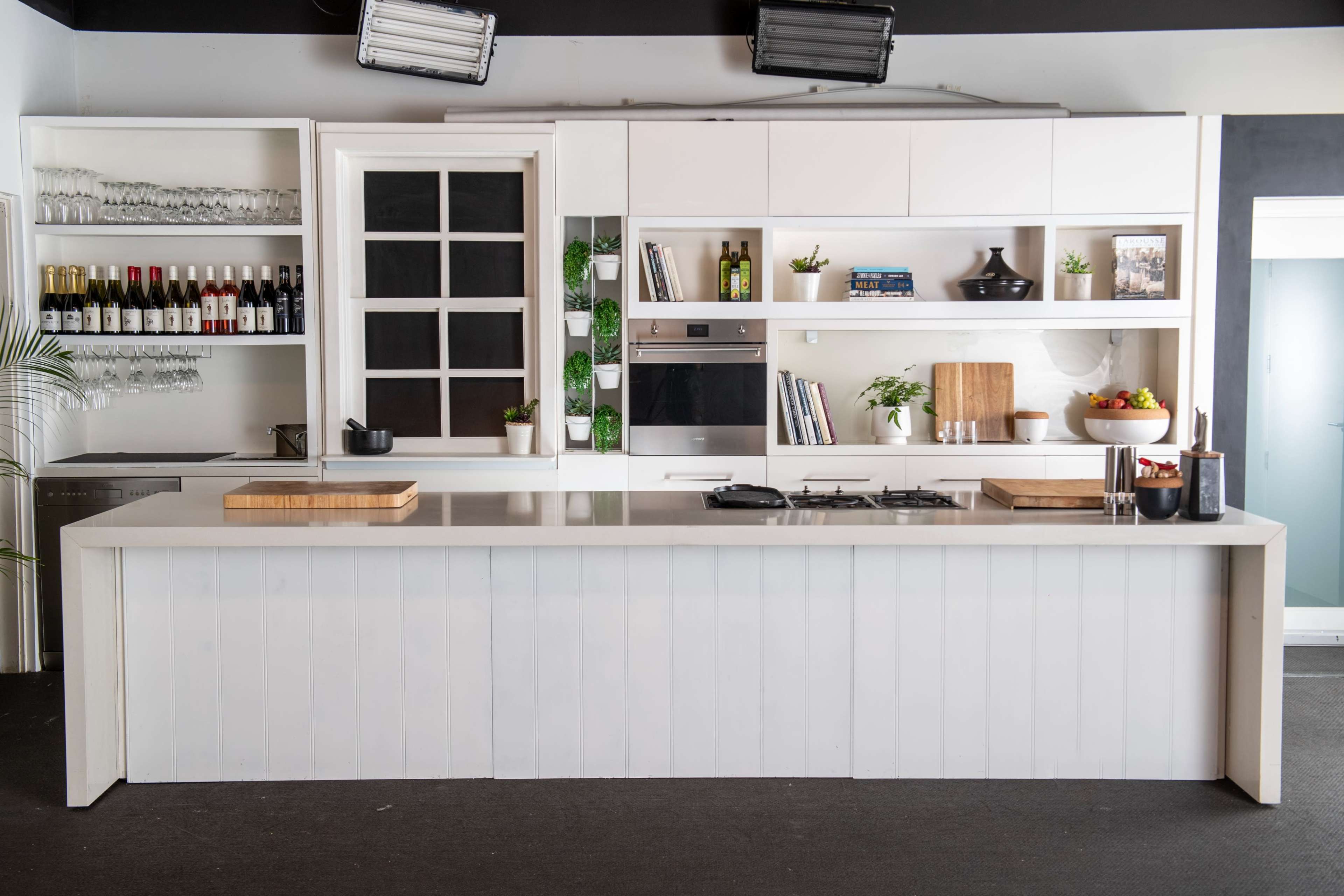 A modern kitchen with a large white island, built-in appliances, shelves holding glassware and bottles, and minimalist decor.