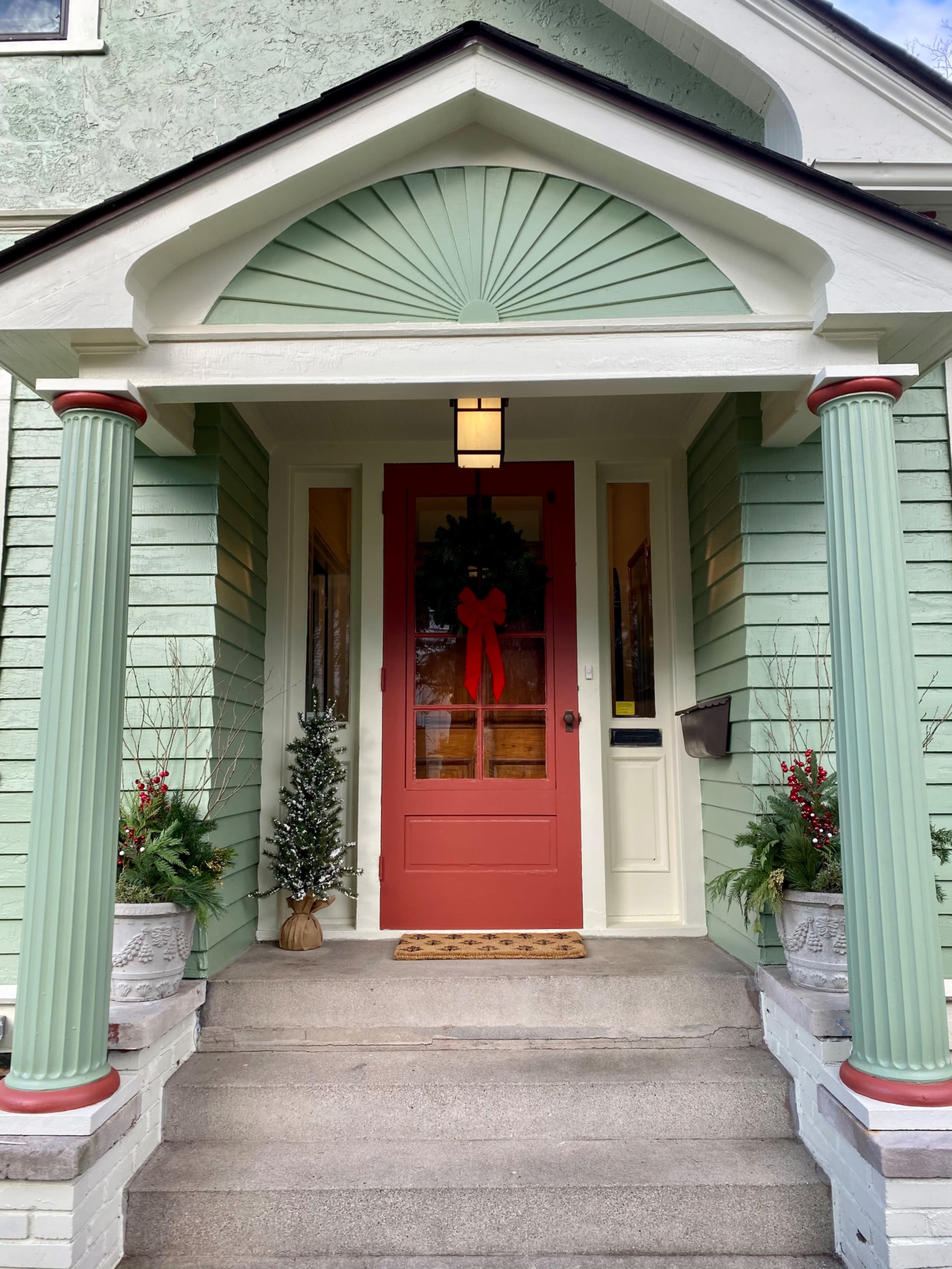 The image shows a house entrance with a red door, green columns, and decorative potted plants on either side.