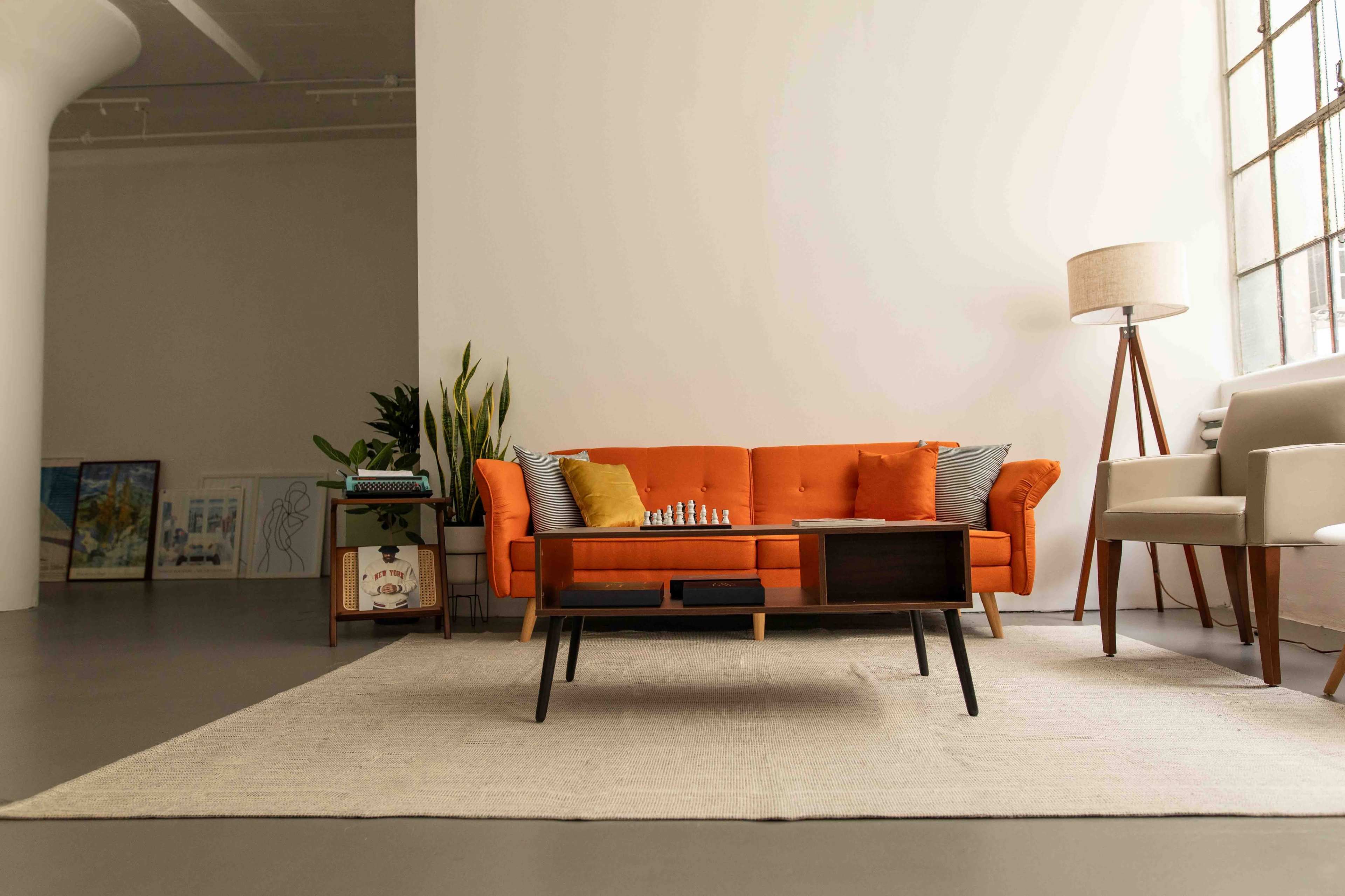 A modern living room with an orange sofa, a coffee table with a chess set, and a pair of plants beside a floor lamp and a gray armchair.