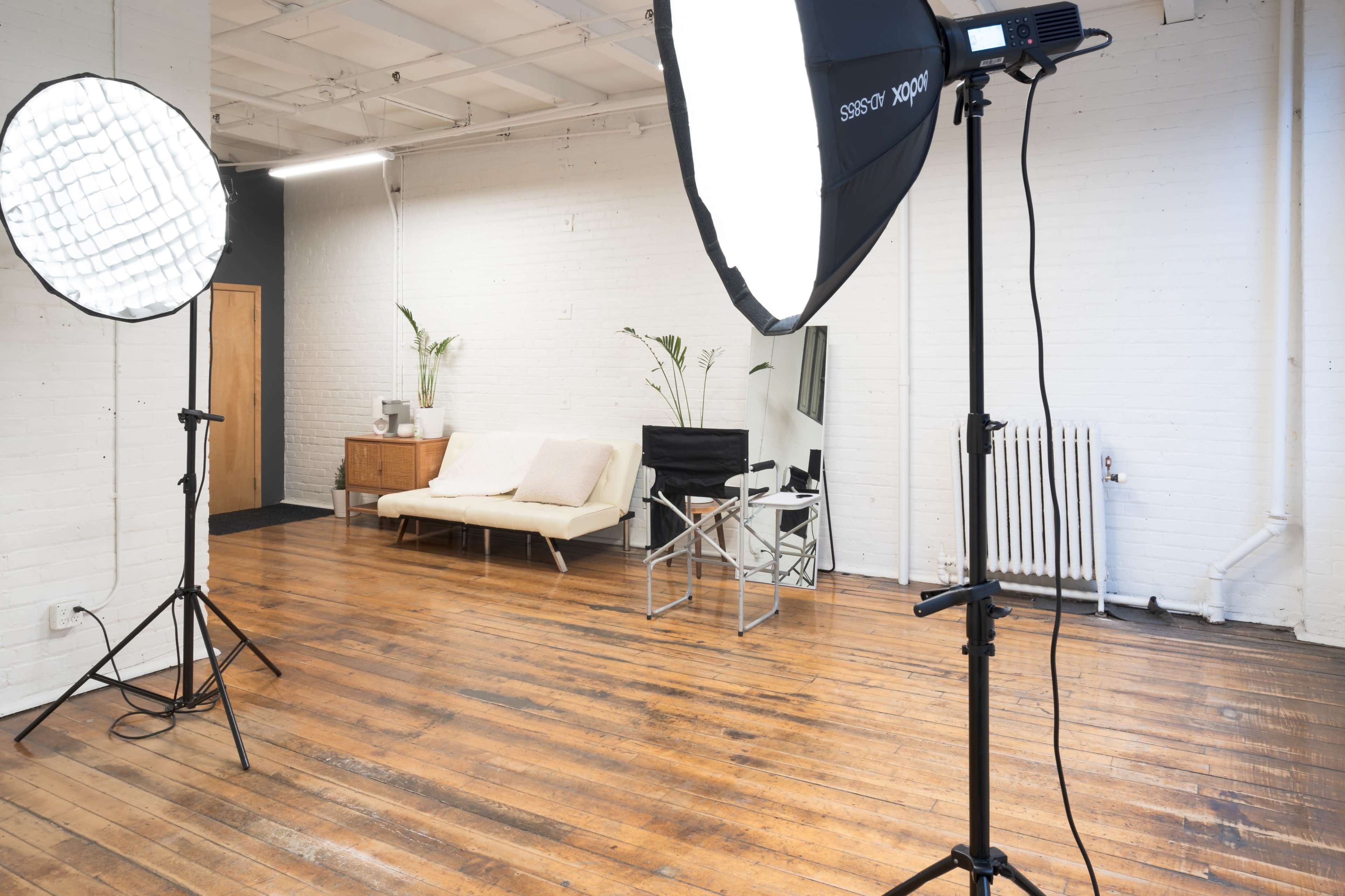 A bright, minimalistic studio space with two lighting stands, a sofa, a director's chair, and hardwood flooring.