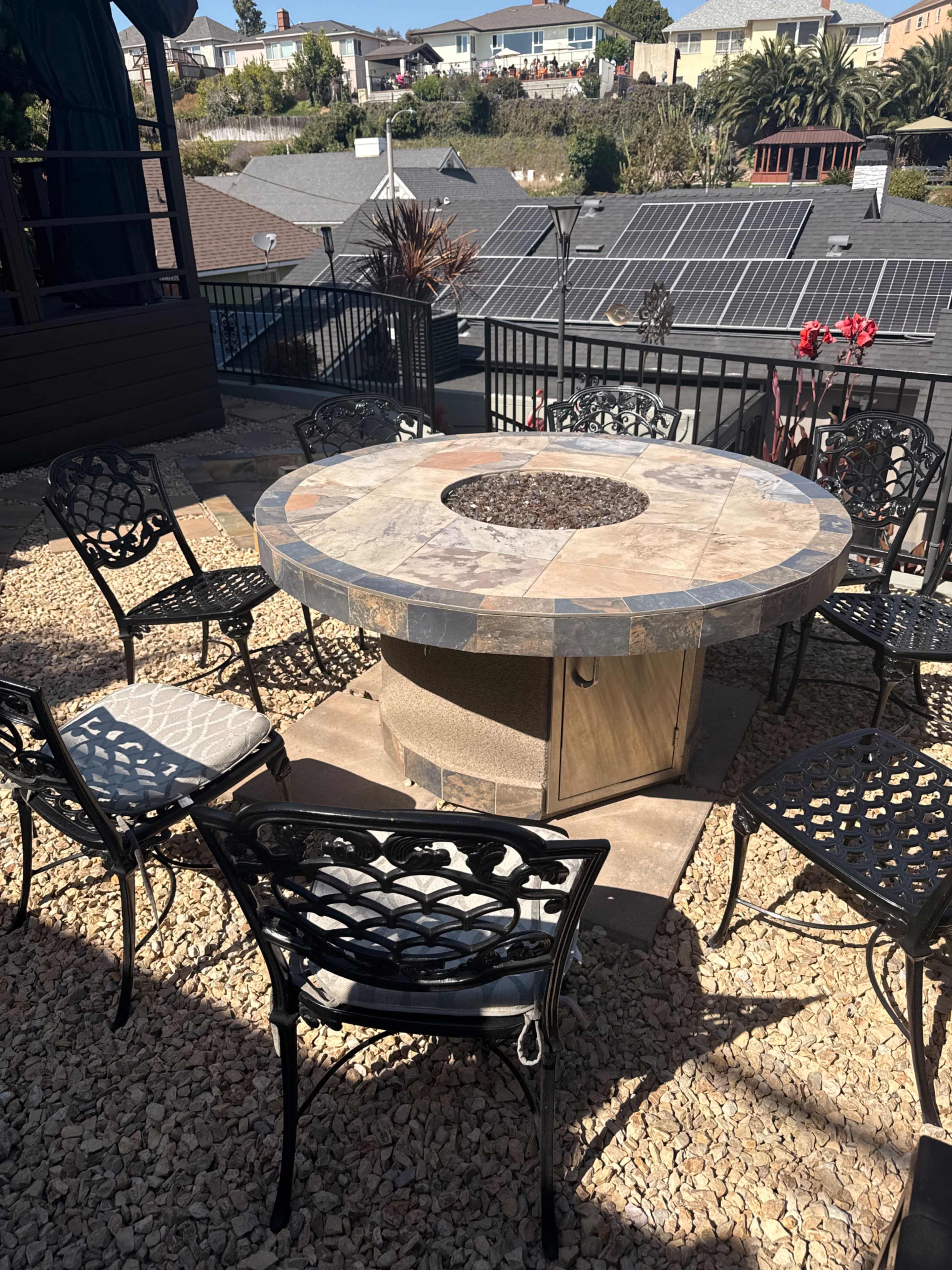 A circular stone fire pit table surrounded by six black metal chairs is situated on a gravel patio with rooftops and solar panels visible in the background.