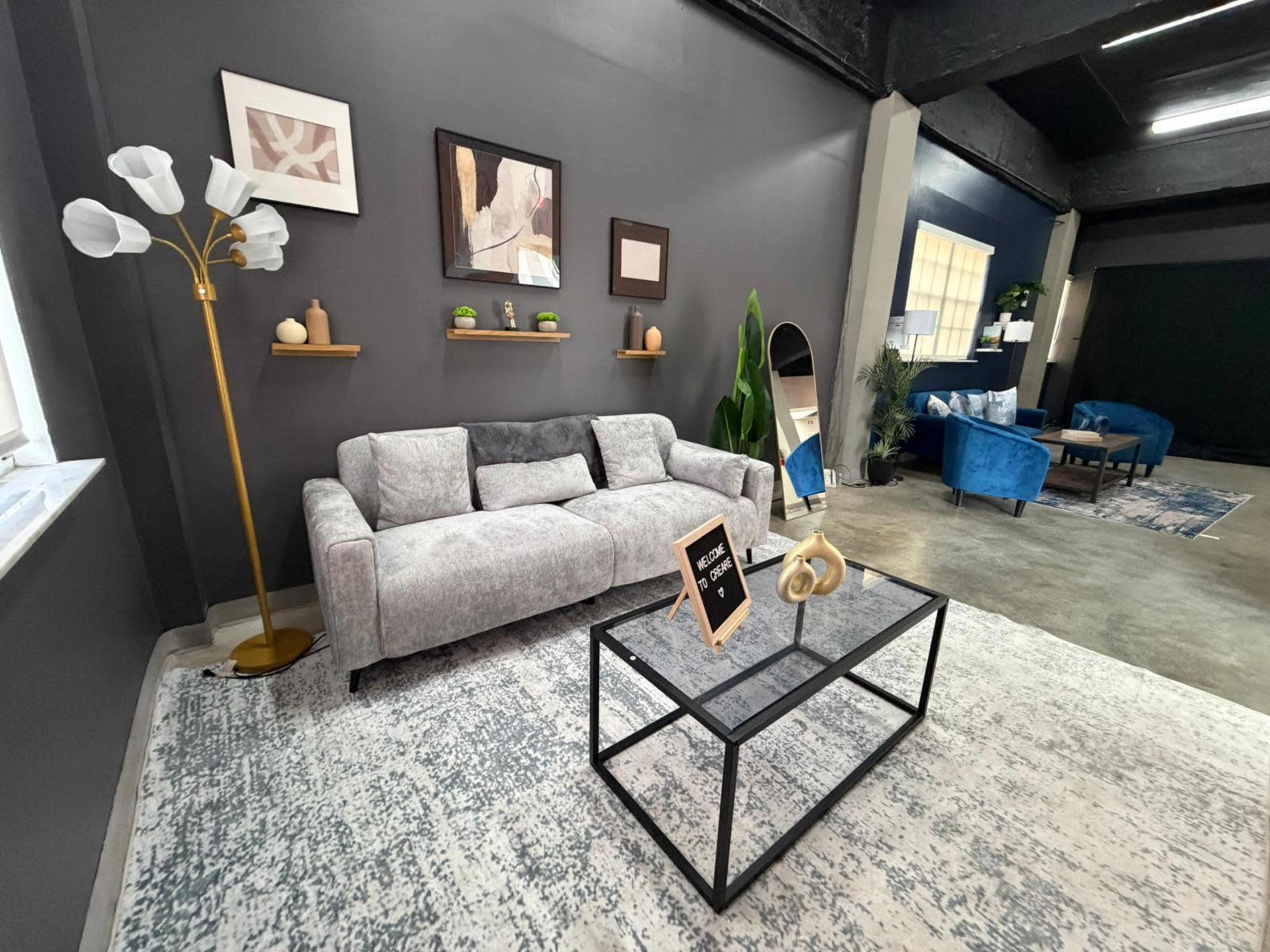 The image shows a modern living space featuring a gray couch, a coffee table, decorative shelves, and blue armchairs against a dark gray wall.