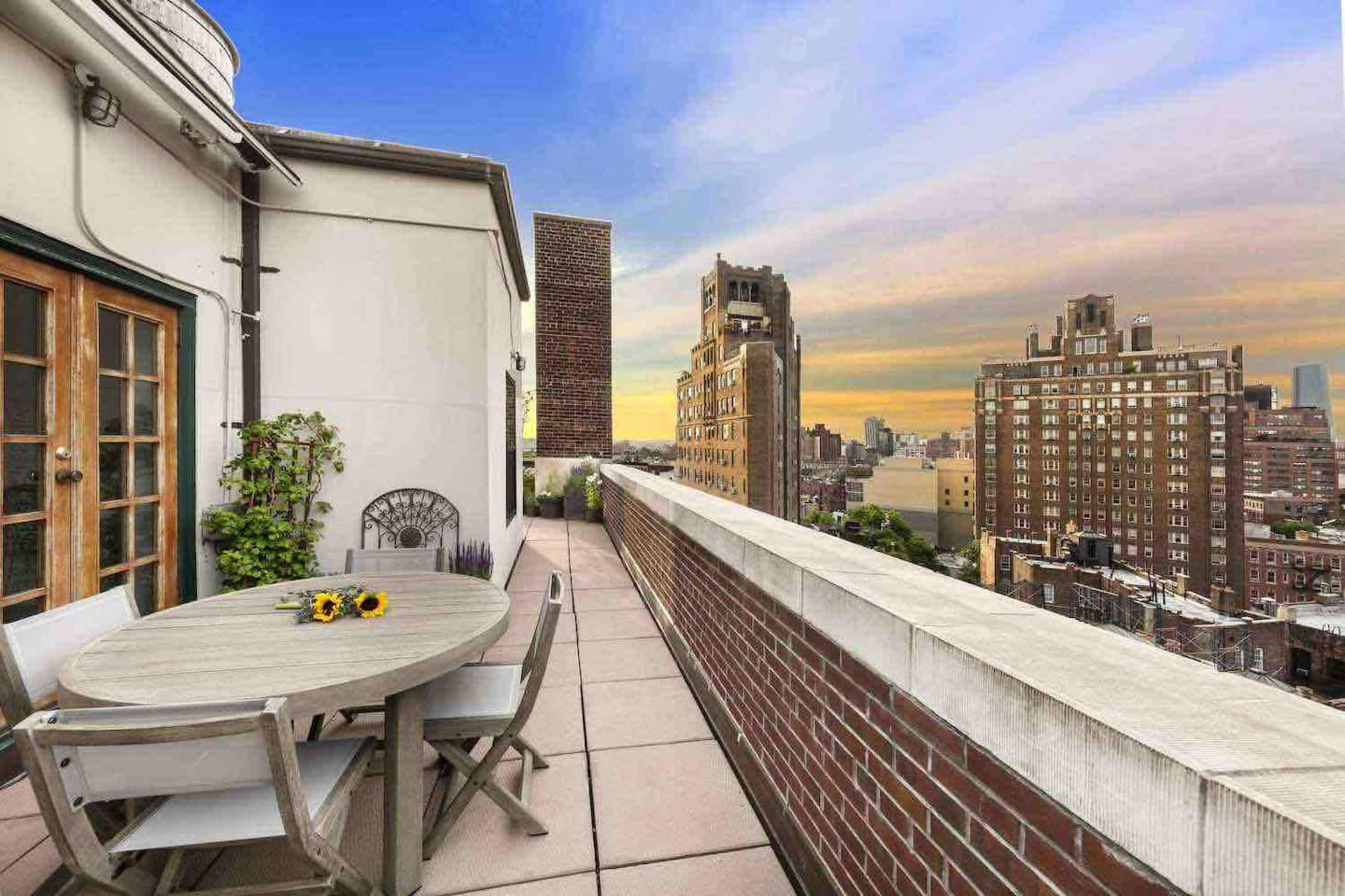 A rooftop terrace features a round table with sunflowers and a view of city buildings against a sunset sky.