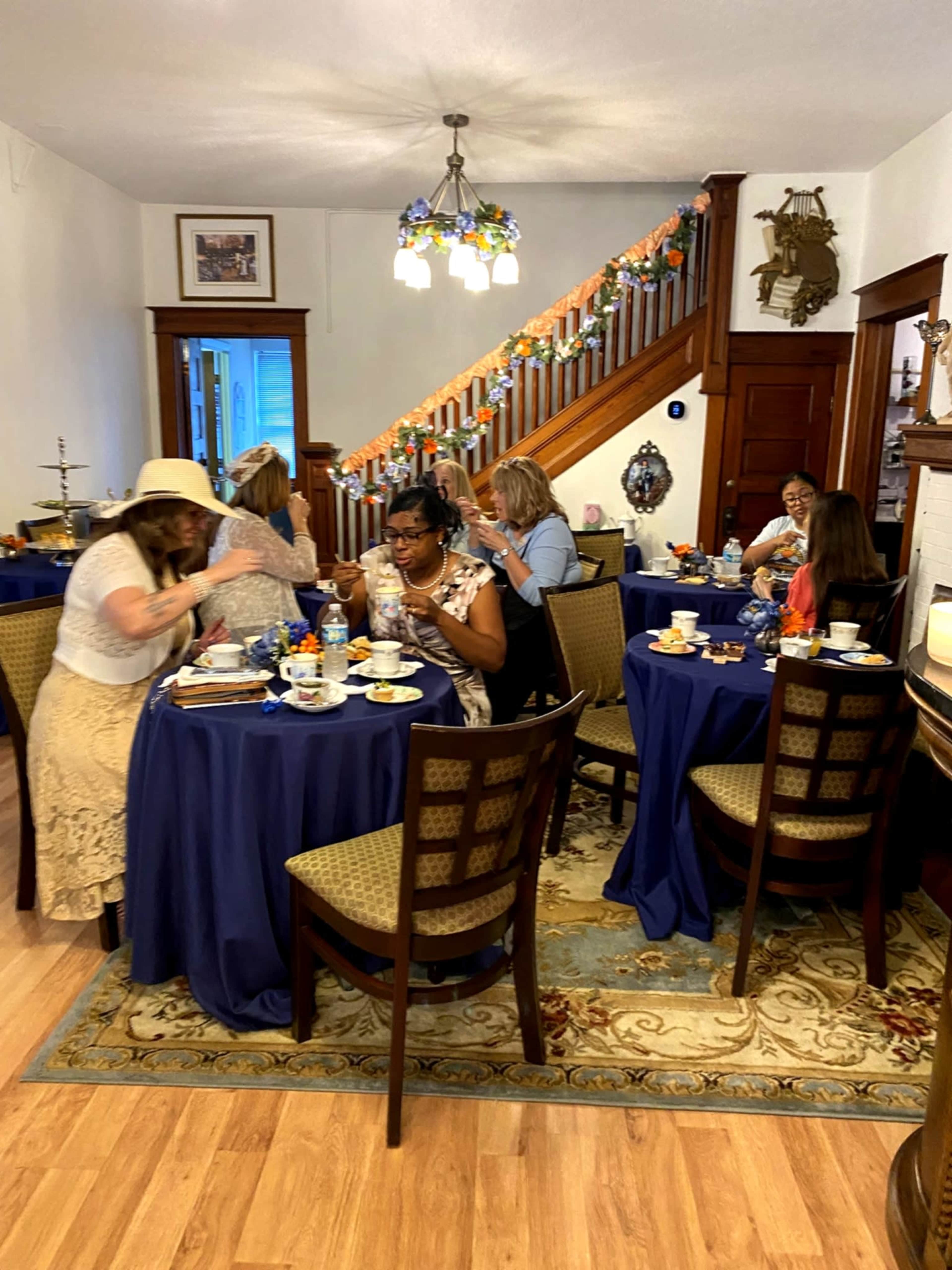 A group of women are seated at elegantly set tables in a cozy dining room, enjoying tea and pastries while some engage in conversation.