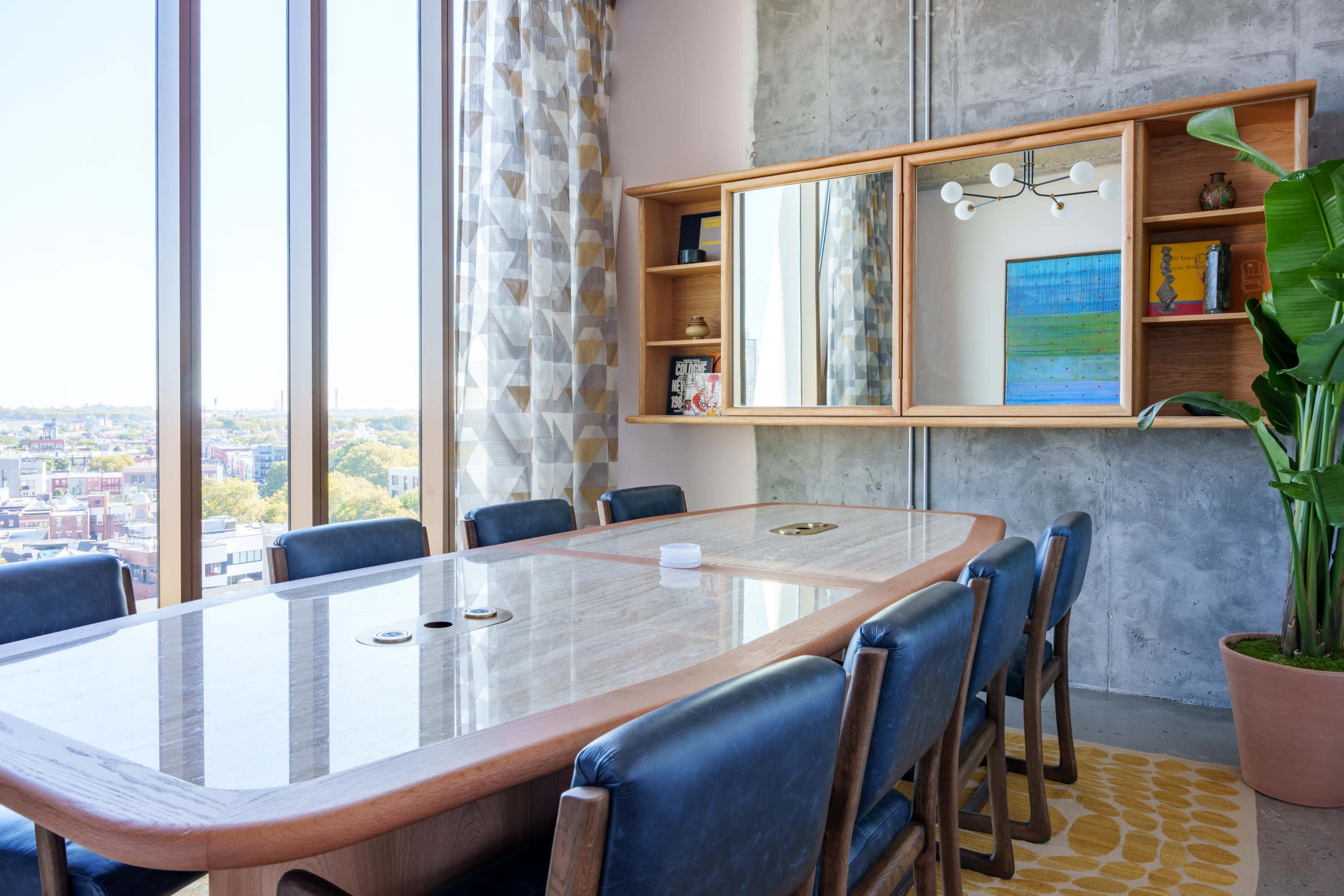 A modern conference room features a large wooden table surrounded by blue chairs, with a mirror and decorative shelving against a concrete wall.