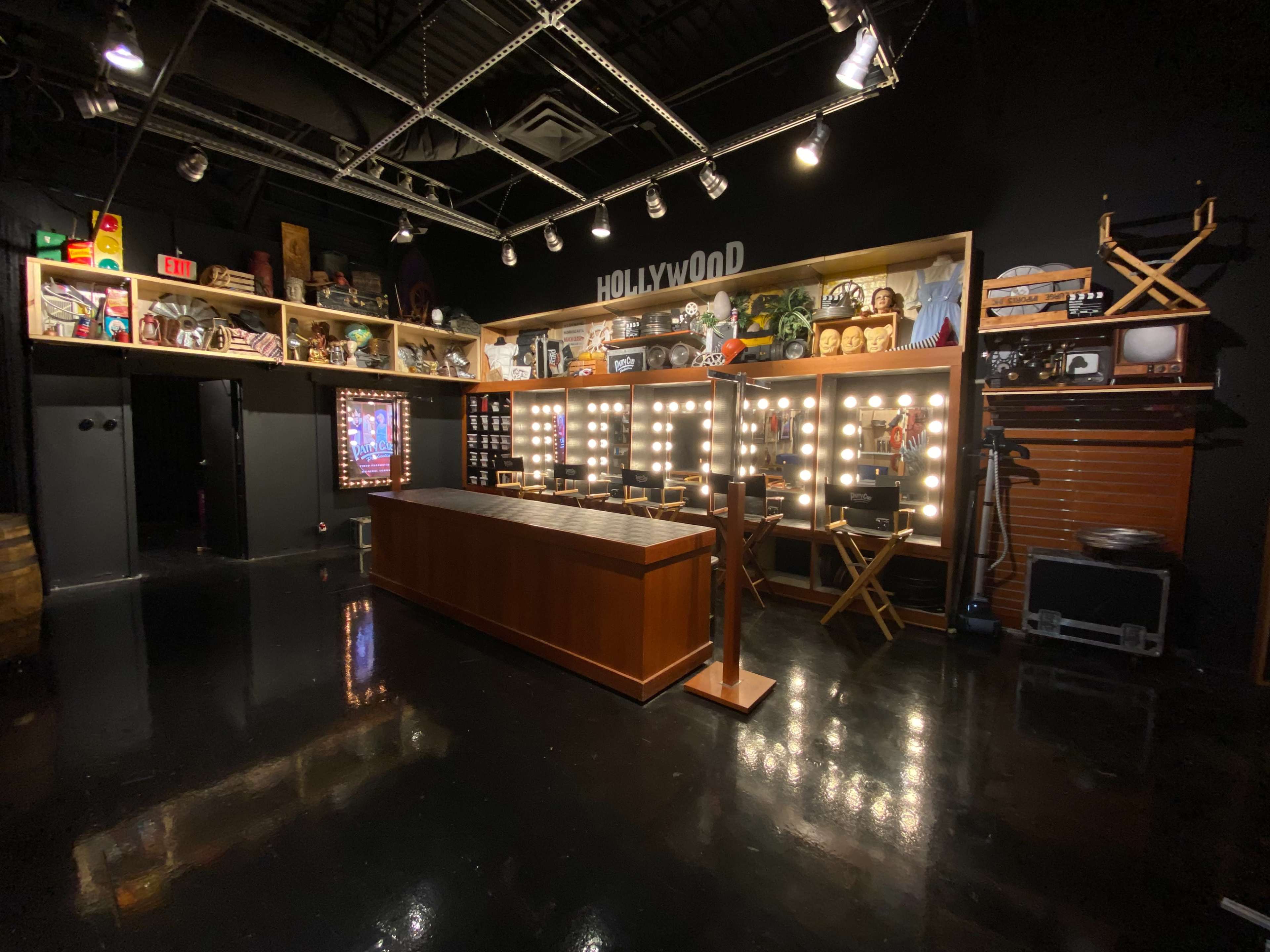 The image shows a Hollywood-themed vanity area featuring a brightly lit mirror setup and a wooden counter, surrounded by shelves displaying various movie memorabilia.