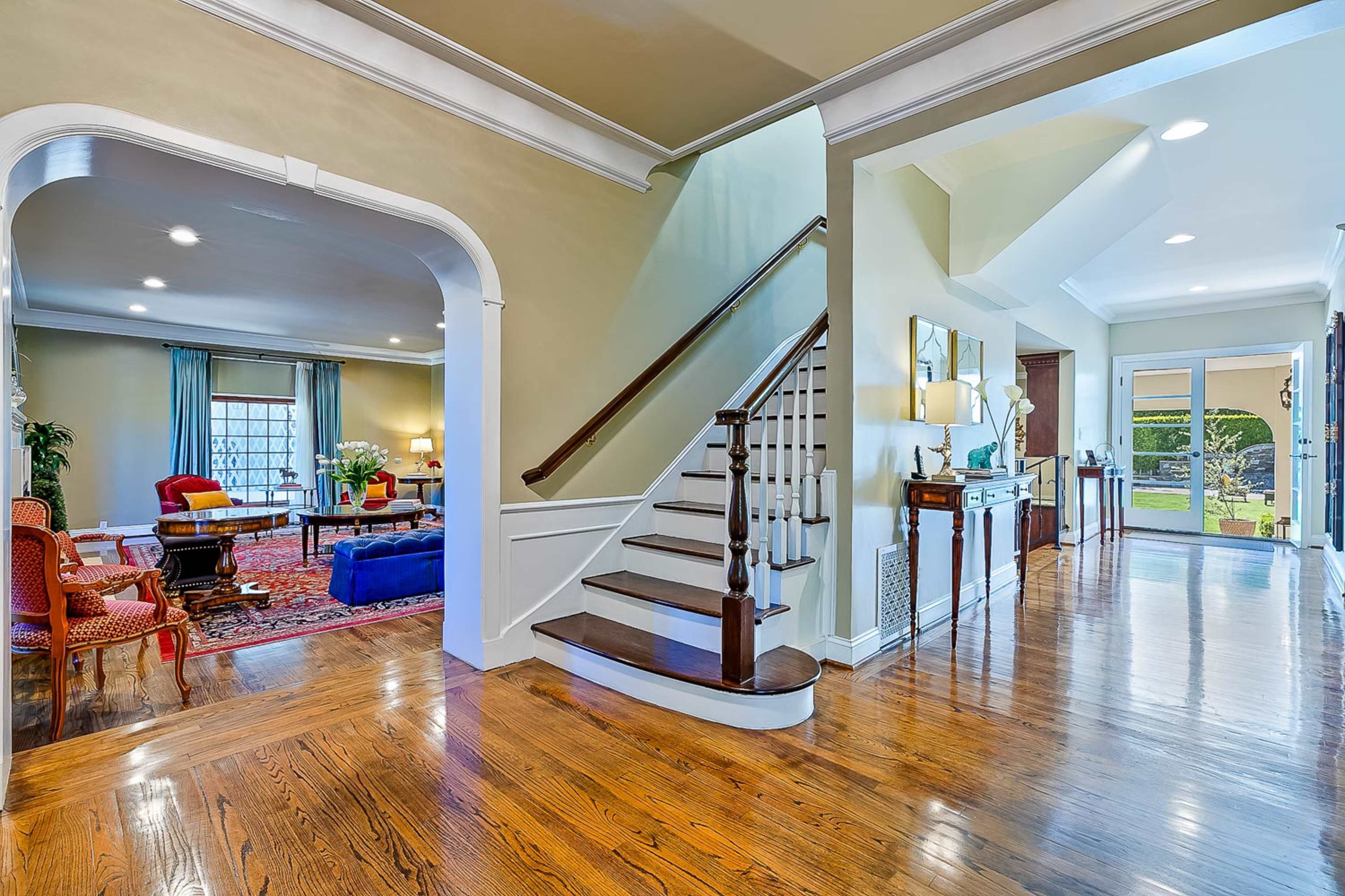 A spacious foyer with a wooden staircase leading to an upper level, and an open archway leading to a living area furnished with a red rug and various seating arrangements.